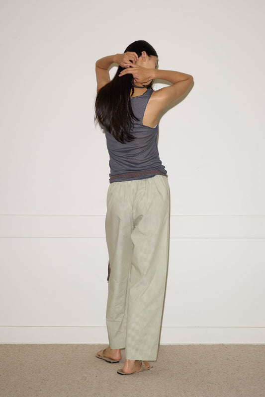 A person with long dark hair wears the Knit Tank - Steel by 0 and loose light pants, standing before a white wall with arms raised—an easy, effortless style in eco-friendly fashion.
