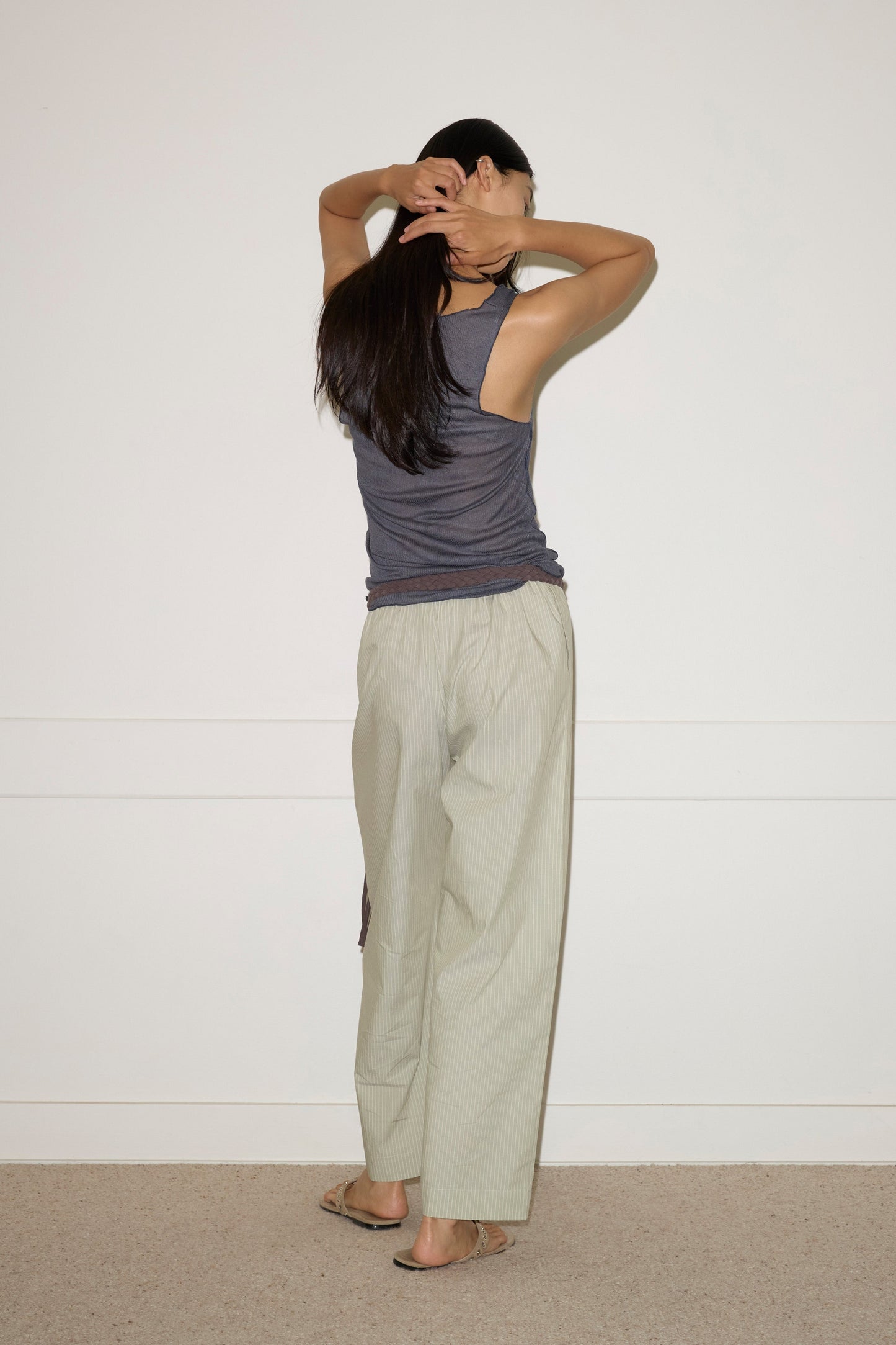 A person with long dark hair stands barefoot against a white wall, wearing a gray sleeveless top and Deiji Studios Ease Trouser - Pistachio Stripe in organic cotton, facing away and adjusting their hair with both hands.