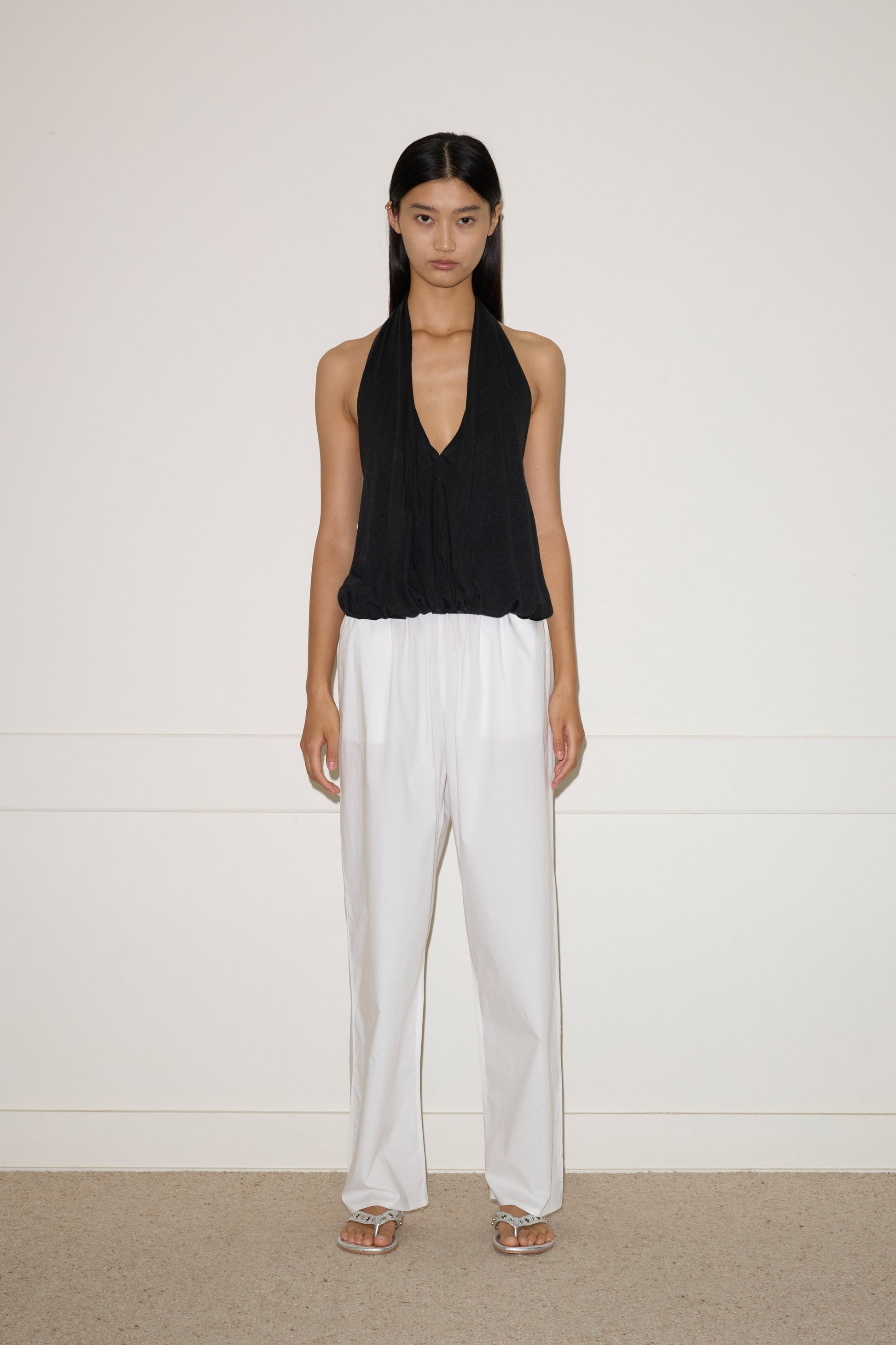 A woman with long dark hair stands against a white wall, wearing the Jersey Top - Black by 0, loose white French linen pants, and white sandals. She has a neutral expression and her arms rest naturally at her sides.