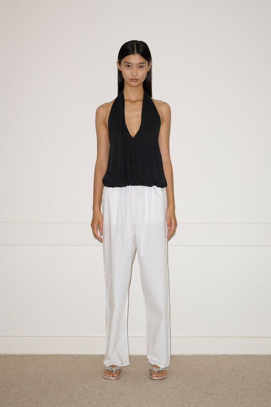 A woman with long dark hair stands against a white wall, wearing the Jersey Top - Black by 0, loose white French linen pants, and white sandals. She has a neutral expression and her arms rest naturally at her sides.