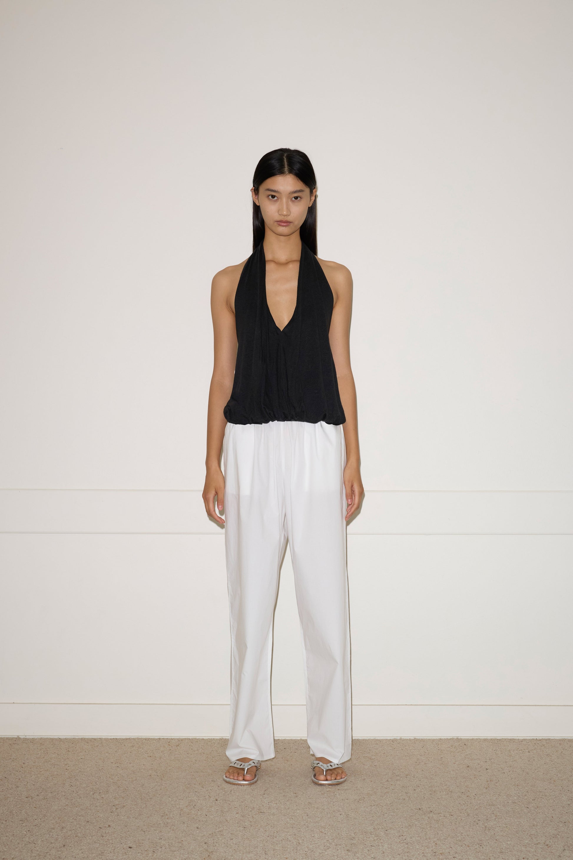 A woman with long dark hair stands against a white wall, wearing the Jersey Top - Black by 0, loose white French linen pants, and white sandals. She has a neutral expression and her arms rest naturally at her sides.