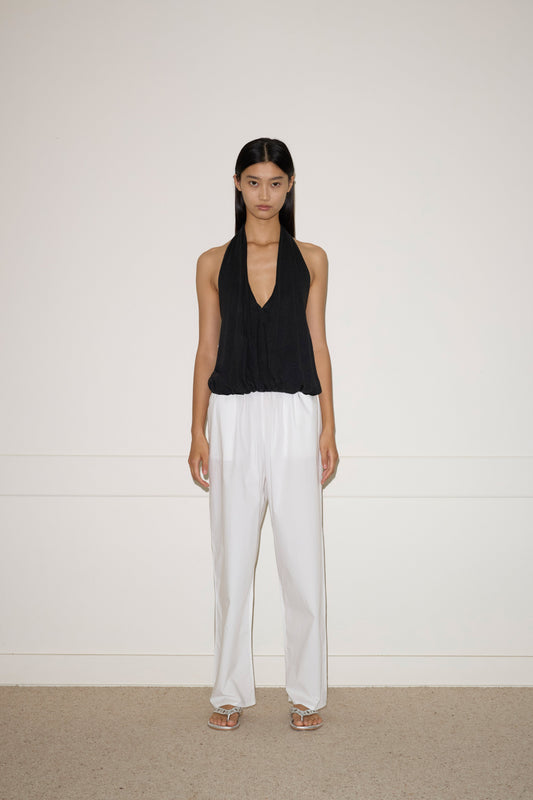 A woman with long dark hair stands against a white wall, wearing the Jersey Top - Black by 0, loose white French linen pants, and white sandals. She has a neutral expression and her arms rest naturally at her sides.