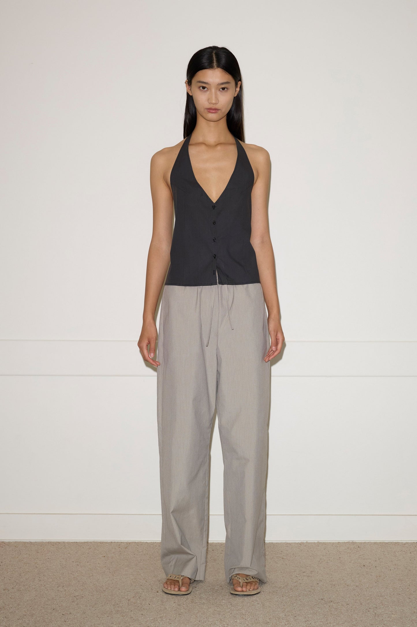 A woman with long, straight dark hair stands barefoot on a beige carpet against a white wall, wearing Deiji Studios Waist Halter - Black Stripe and loose light gray trousers.