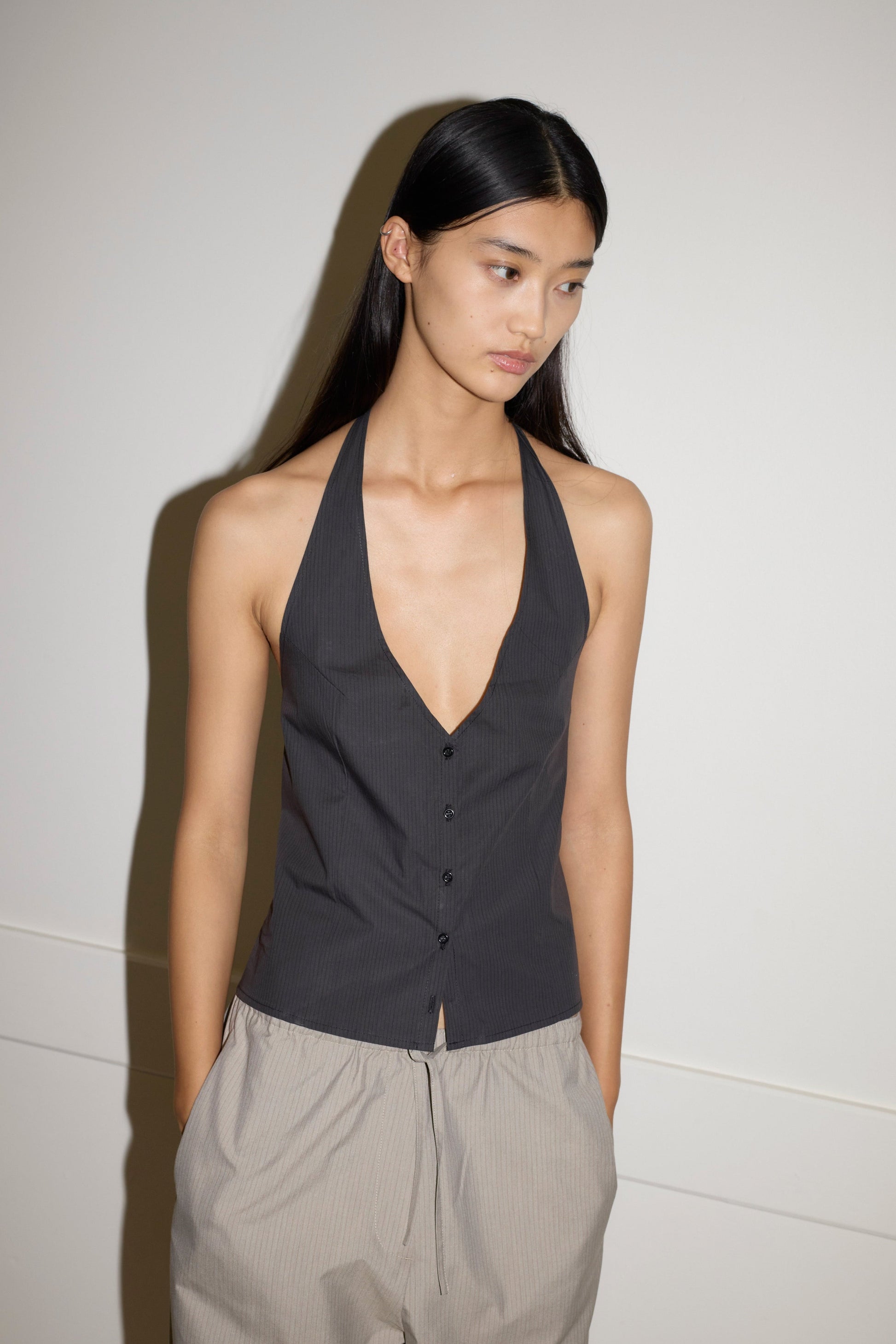 A young woman with long dark hair stands against a plain wall, wearing the Waist Halter top in dark gray and loose light gray pants, her hands in her pockets as she looks slightly away from the camera.
