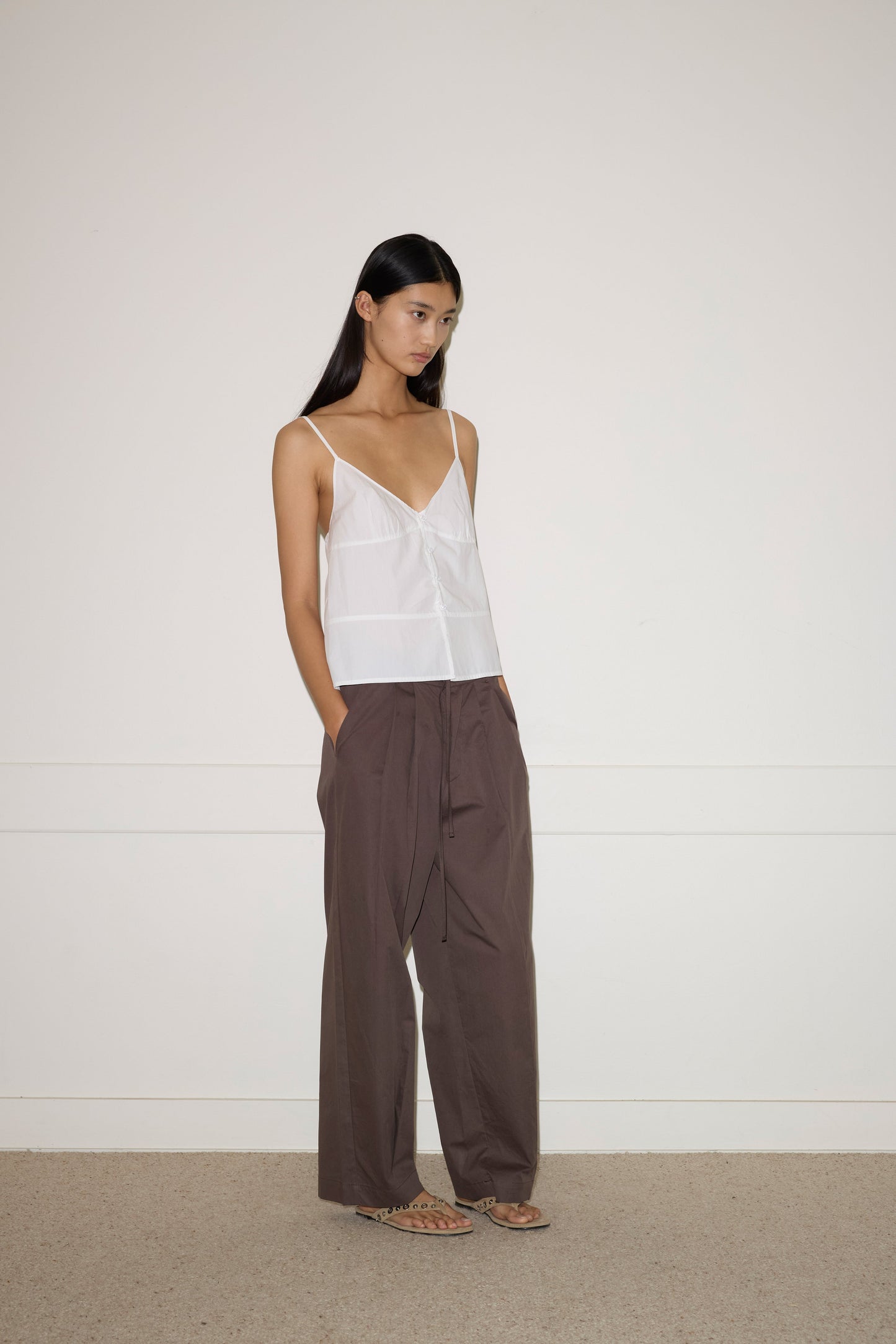 A woman stands against a plain white wall wearing the Seam Top - White by 0—a sleeveless top with thin straps and a subtle button front—paired with loose, high-waisted brown trousers. Her long straight hair falls as her hands rest in her pockets.