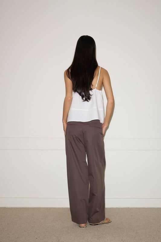 A woman with long dark hair stands facing a white wall, wearing a white sleeveless top and the Turn Pant - Cocoa by 0. Her hands are in her pockets and her back is to the camera.