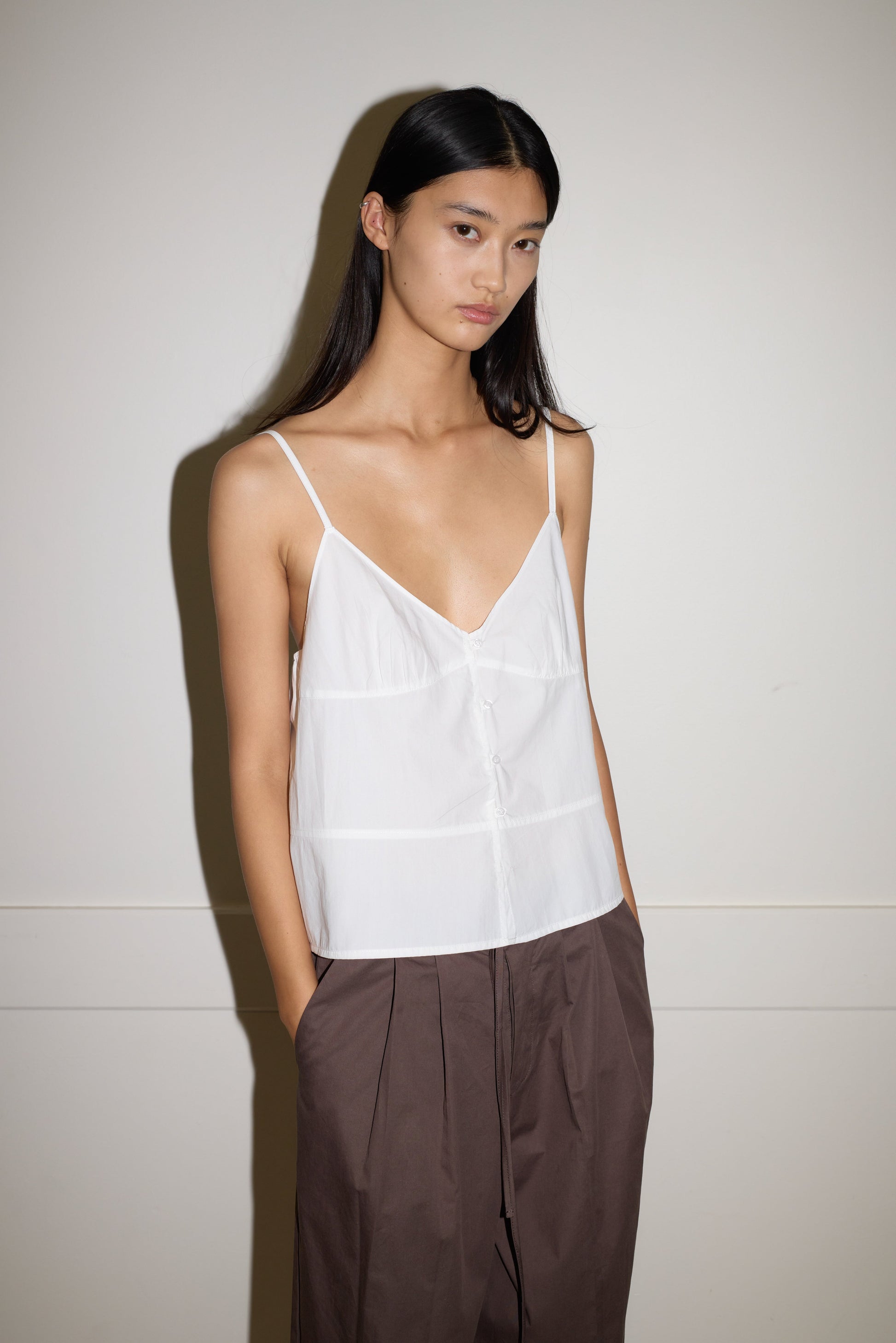 A person with long dark hair wears the Seam Top - White by 0, a white organic cotton spaghetti strap top, and brown wide-leg pants, standing against a plain light-colored wall with a neutral expression.