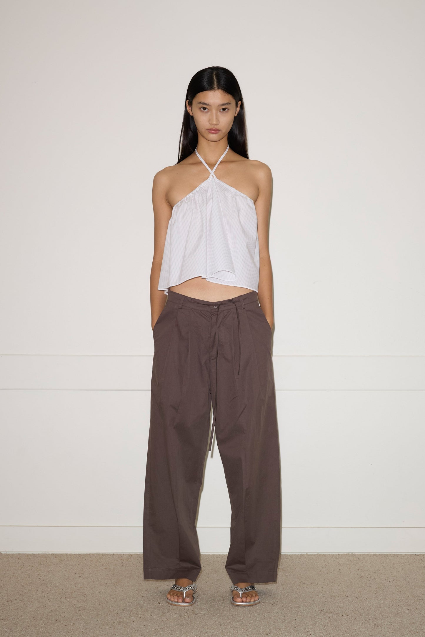 A woman stands against a plain wall wearing the 0 Napkin Top - Pacific Stripe, loose brown tie-waist pants, and flat sandals. Her hair is straight and she faces the camera with a neutral expression.