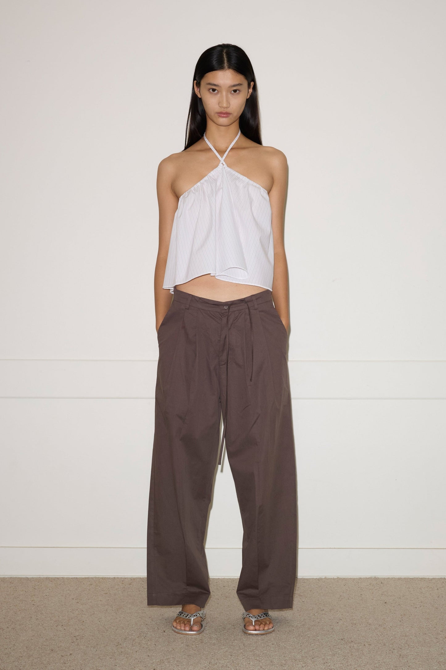 A woman stands against a plain wall, wearing the Turn Pant - Cocoa by 0, featuring relaxed tailoring, organic cotton fabric, and an adjustable drawcord waist. She pairs them with a white halter-neck top and silver sandals. Her hair is straight; her expression neutral.