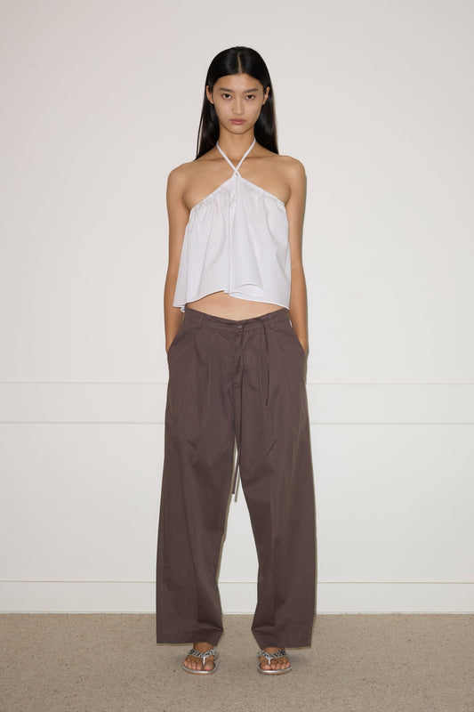 A woman stands against a plain wall, wearing the Turn Pant - Cocoa by 0, featuring relaxed tailoring, organic cotton fabric, and an adjustable drawcord waist. She pairs them with a white halter-neck top and silver sandals. Her hair is straight; her expression neutral.