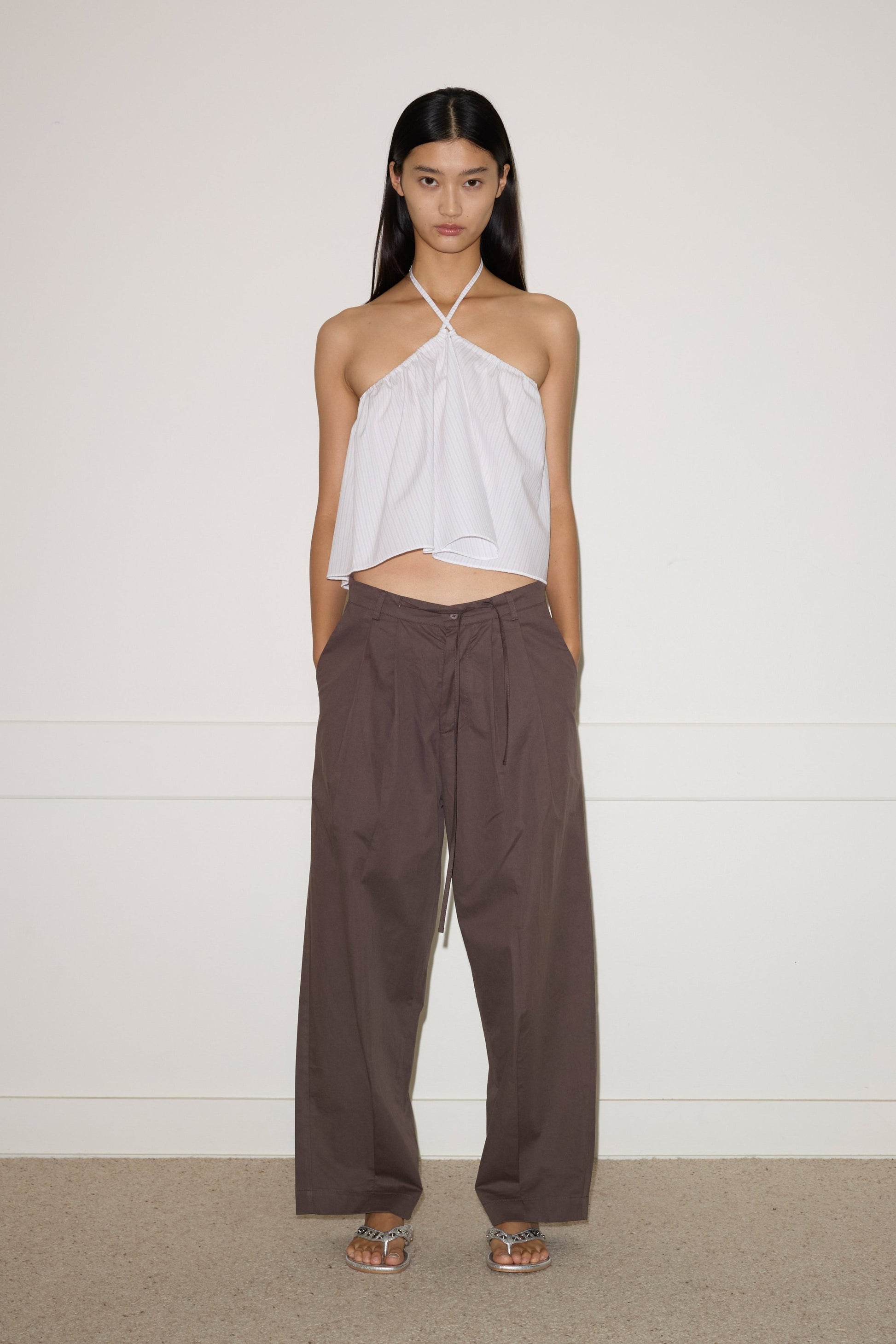 A woman with long, straight dark hair stands against a plain wall, hands in her pockets, wearing the Turn Pant—a loose-fitting brown tie-waist style—with a sleeveless white halter top and sandals.