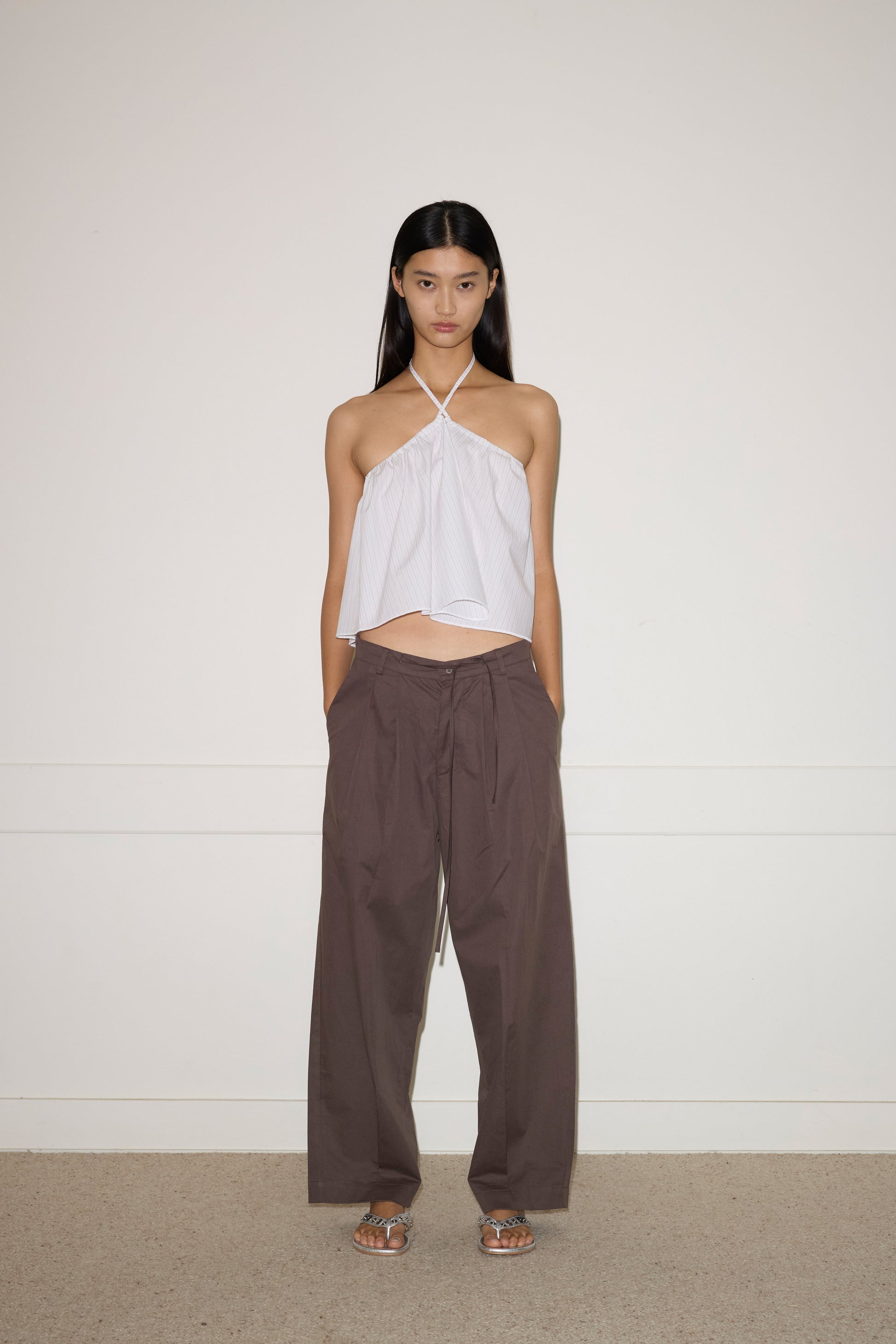 A woman stands against a plain wall, wearing the Turn Pant - Cocoa by 0, featuring relaxed tailoring, organic cotton fabric, and an adjustable drawcord waist. She pairs them with a white halter-neck top and silver sandals. Her hair is straight; her expression neutral.