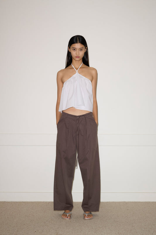 A woman stands against a plain wall, wearing the Turn Pant - Cocoa by 0, featuring relaxed tailoring, organic cotton fabric, and an adjustable drawcord waist. She pairs them with a white halter-neck top and silver sandals. Her hair is straight; her expression neutral.