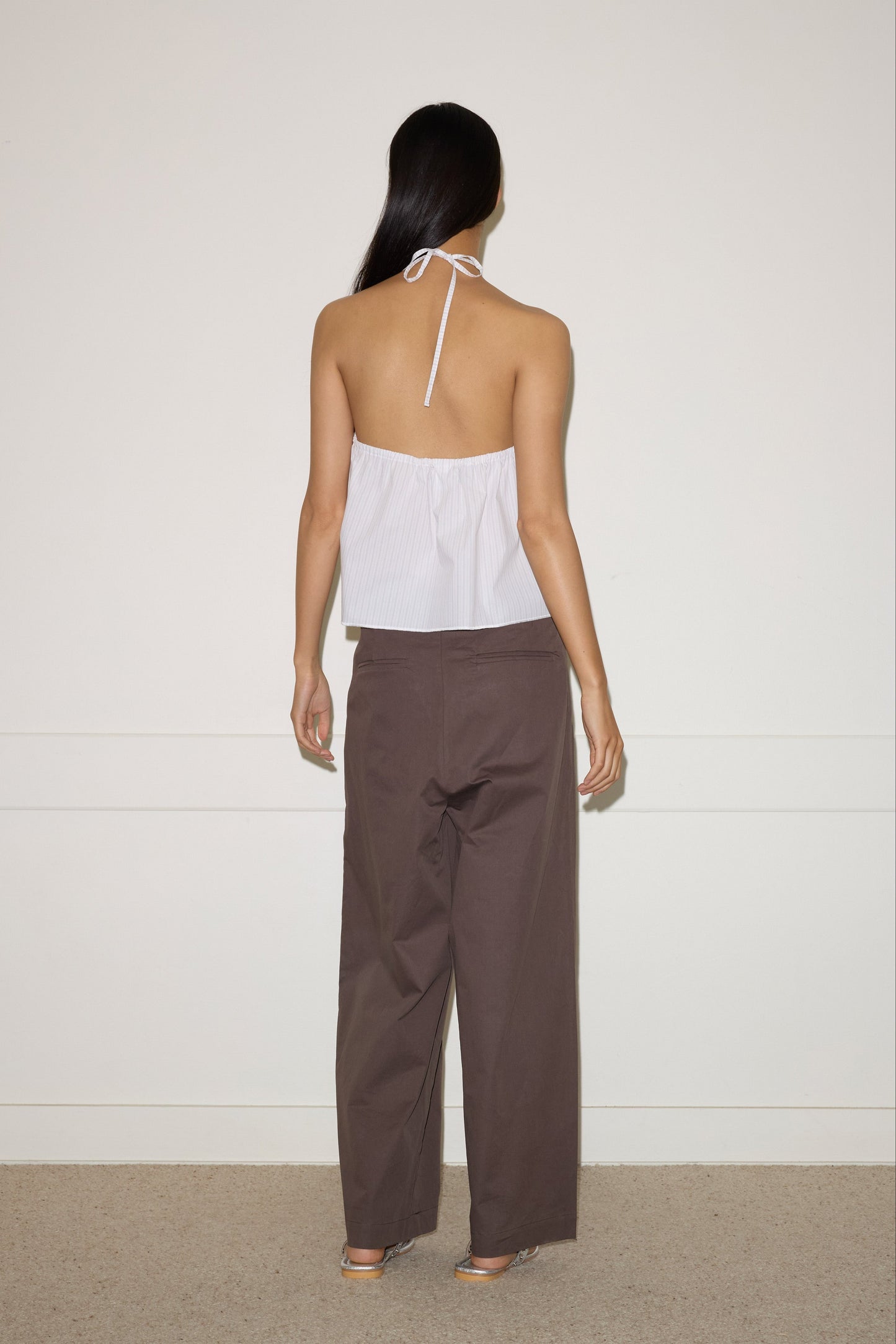 A person with long dark hair stands facing a plain white wall, wearing the Napkin Top - Pacific Stripe from 0 and loose brown pants. Their arms are relaxed at their sides, and their bare feet rest on a beige carpeted floor.