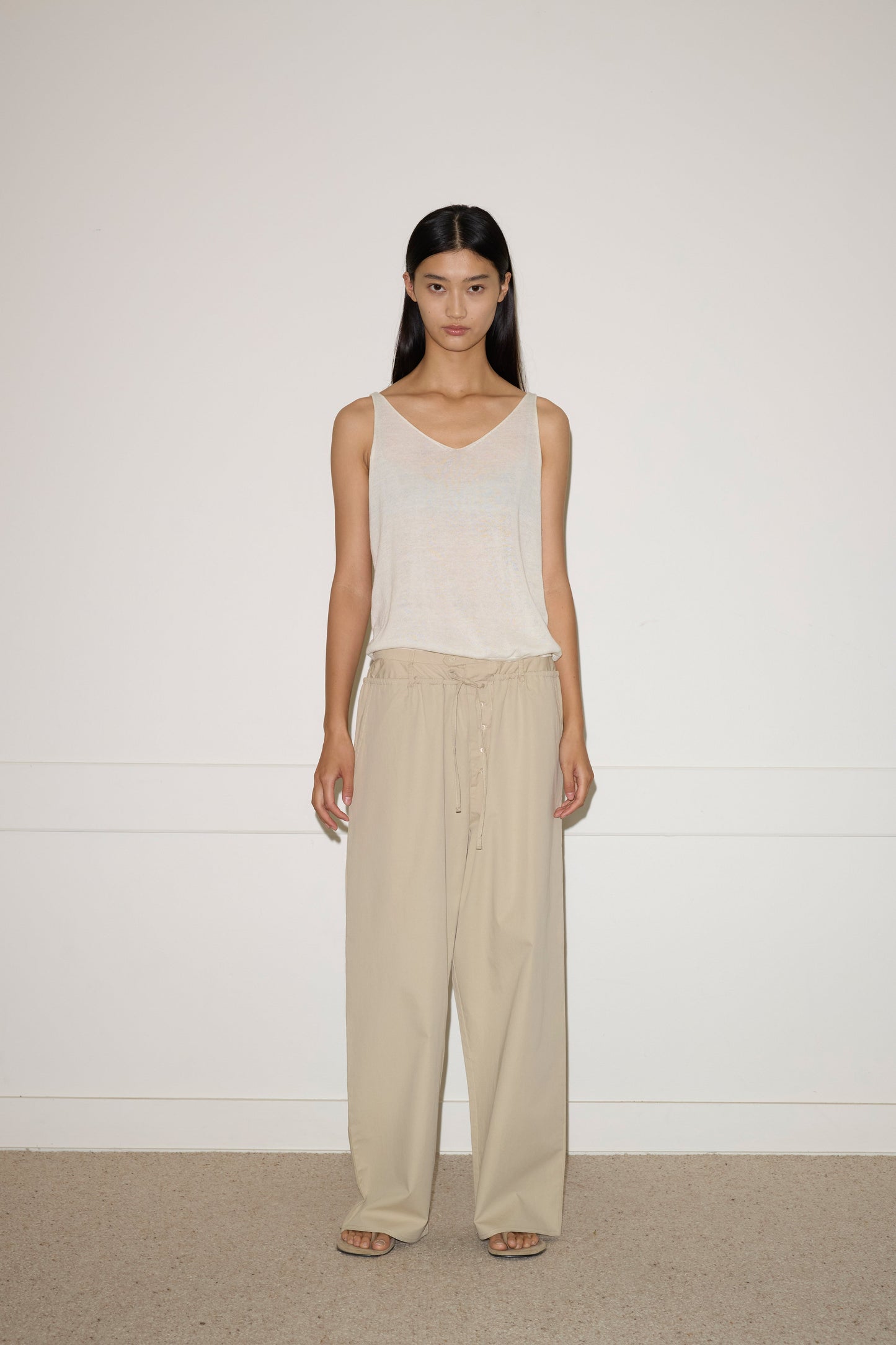 A person stands against a plain white wall, wearing the 0 Rolled Knit Top in Off White, loose beige drawstring pants, and beige sandals. They have long, straight dark hair and a neutral expression.