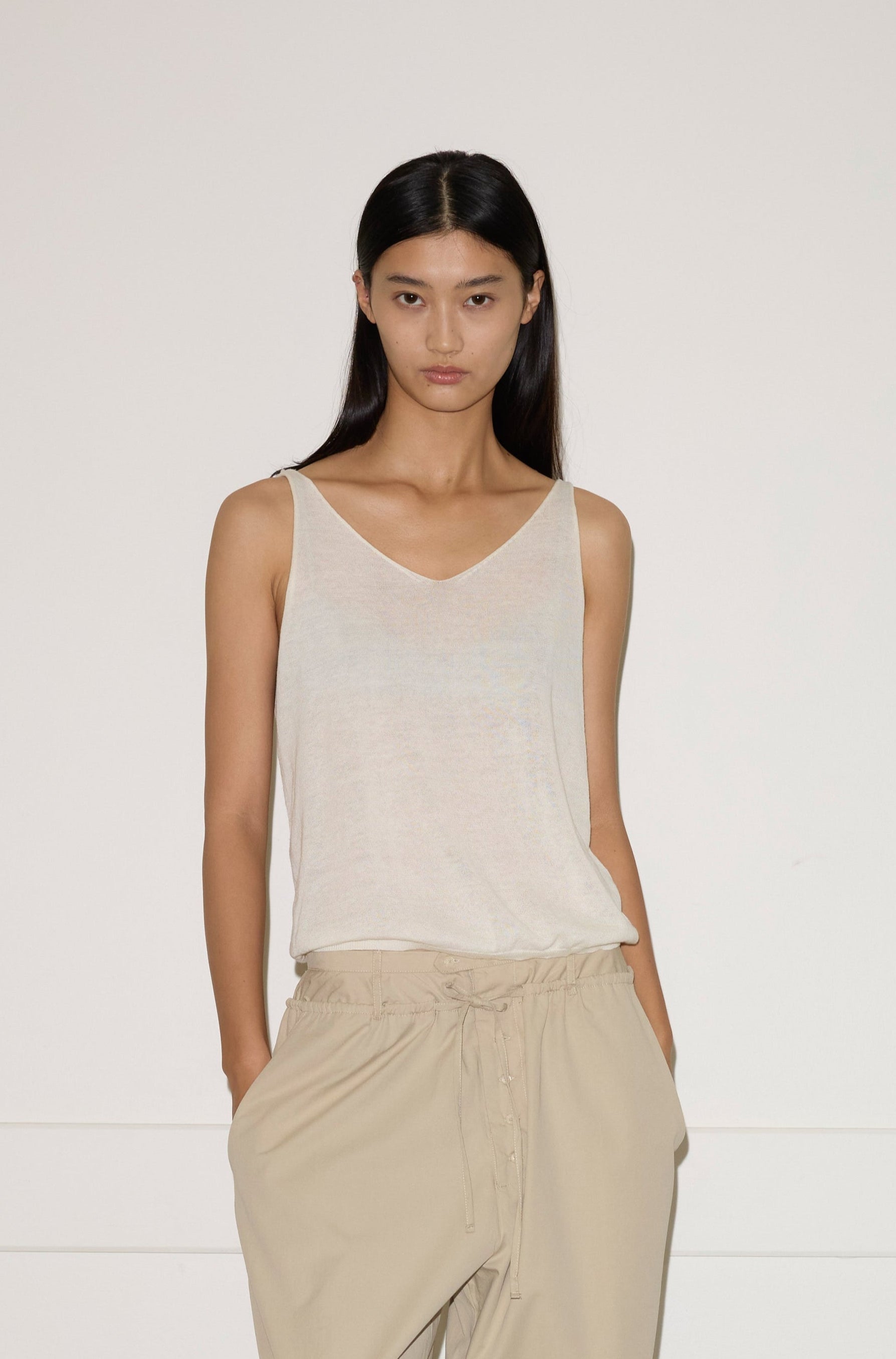 A woman stands against a white wall wearing the 0 Rolled Knit Top in off-white with loose beige pants, her hands in her pockets. She has straight dark hair and a neutral expression.