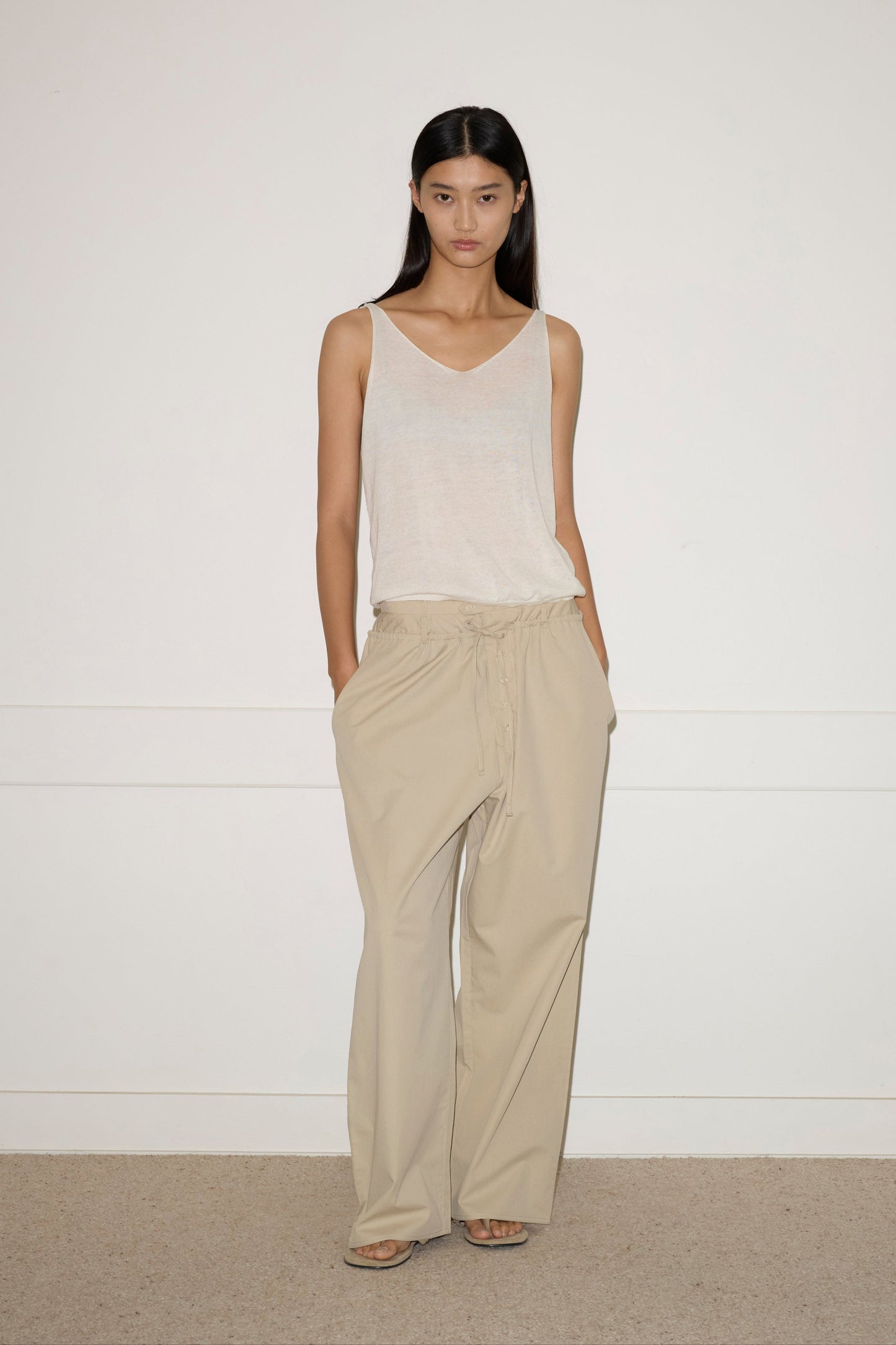 A woman with long dark hair and a neutral expression stands indoors against a plain white wall, wearing the Two Band Pant - Fawn in organic cotton by Deiji Studios and a sheer white sleeveless top, her hands casually in her pockets.