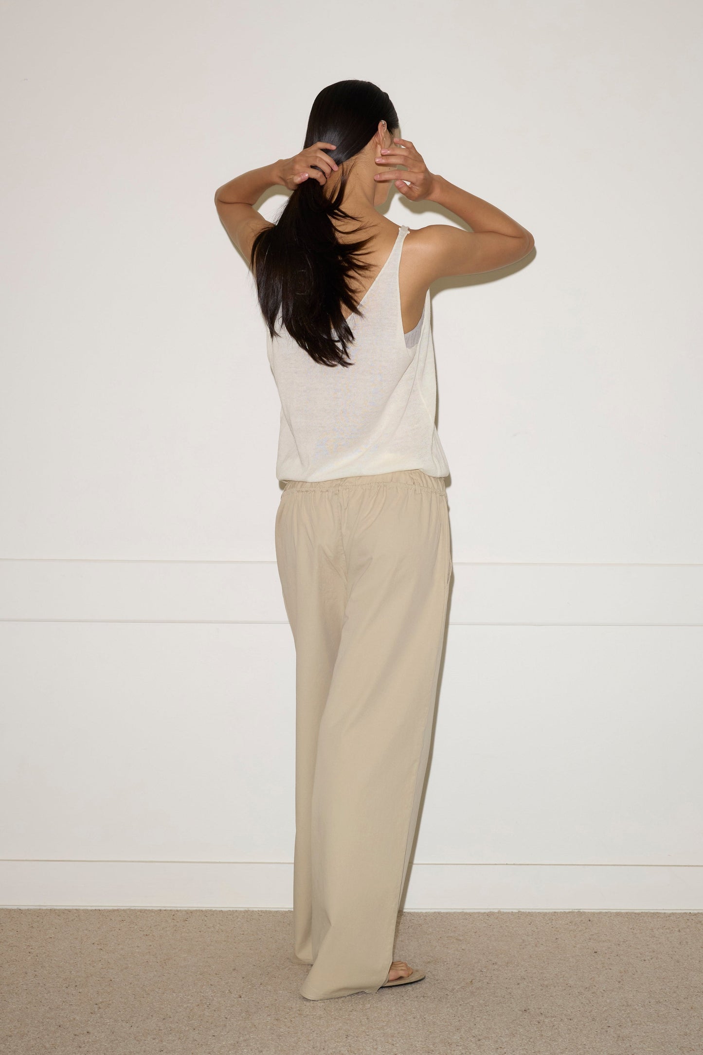 A woman stands against a white wall, facing away from the camera in a sleeveless top and the Two Band Pant - Fawn by Deiji Studios, crafted from breathable organic cotton for an effortlessly chic look.