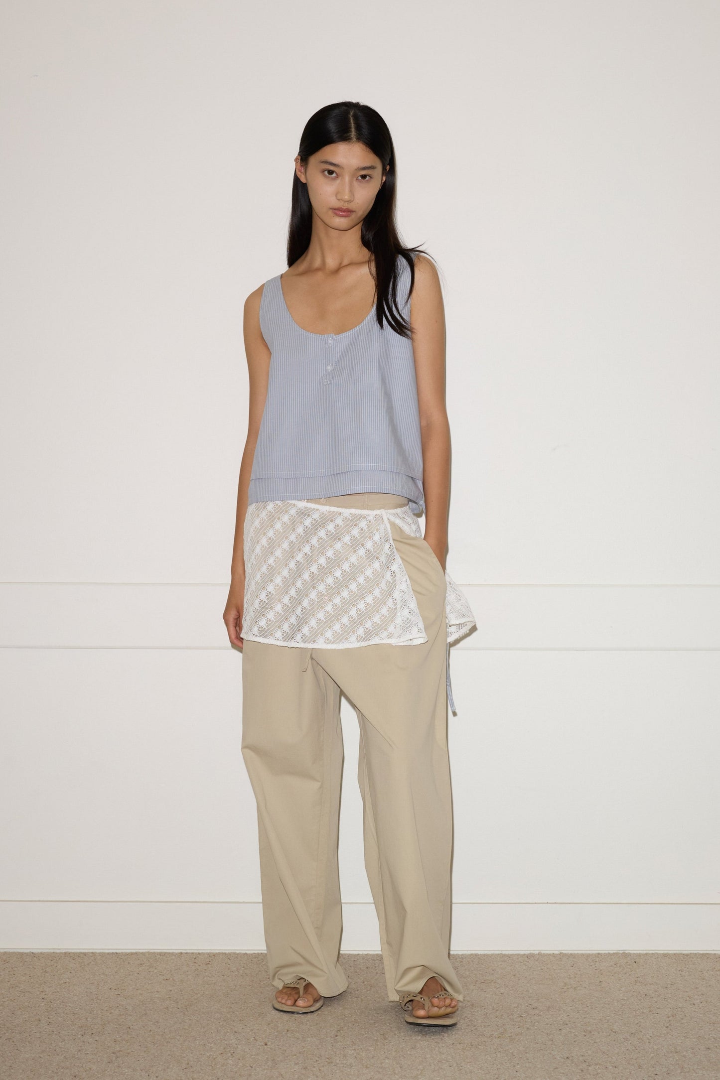 A woman stands against a white wall, wearing the 0 Layered Top - Sky Stripe, loose beige pants, tan sandals, and a white patterned apron tied at her waist.