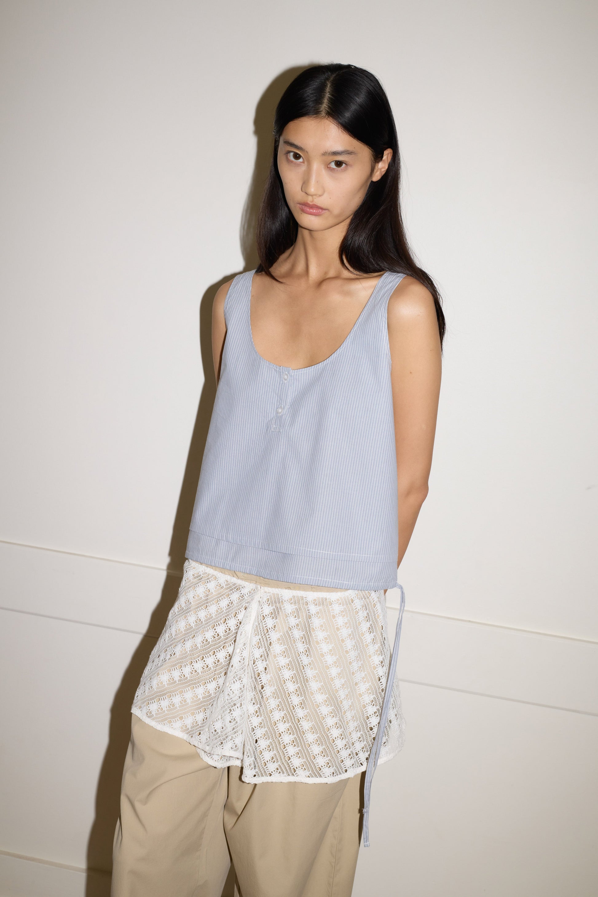 A woman with long dark hair stands against a plain wall, wearing the 0 Layered Top - Sky Stripe over a white lace shirt and beige wide-leg pants—her expression neutral.