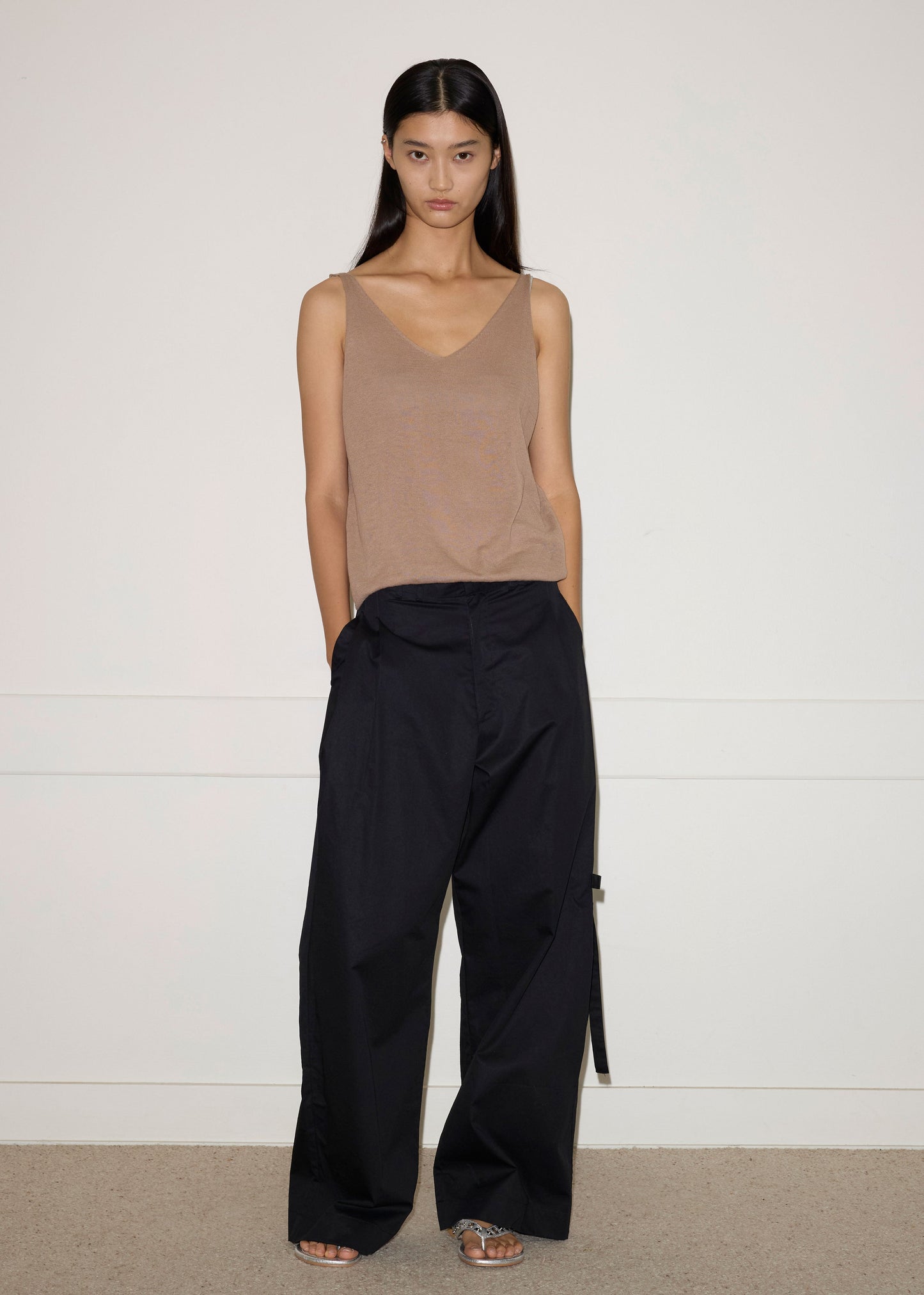 A woman stands against a plain white wall, wearing a tan sleeveless top and the Tab Pant - Black by Deiji Studios. Her hands are in her pockets and she has long straight dark hair with a neutral expression.