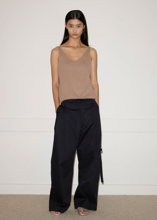A woman stands against a plain white wall, wearing a tan sleeveless top and the Tab Pant - Black by Deiji Studios. Her hands are in her pockets and she has long straight dark hair with a neutral expression.