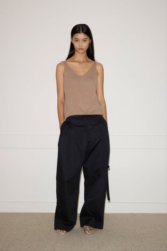 A woman with long dark hair stands against a white wall, wearing the 0 Rolled Knit Top in light brown, loose black pants, and open-toed shoes. She looks directly at the camera with a neutral expression.