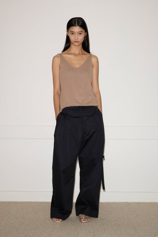 A woman with long dark hair stands against a plain wall, wearing the Tab Pant - Black from 0. She pairs it with a beige sleeveless top and open-toed sandals, resting her hands in her pockets while maintaining a neutral expression.