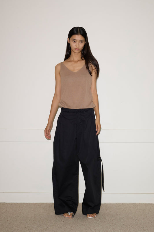 A woman with long dark hair stands indoors against a plain white wall, wearing the 0 Rolled Knit Top in light brown, wide-leg black pants, and sandals. She faces the camera with a neutral expression.