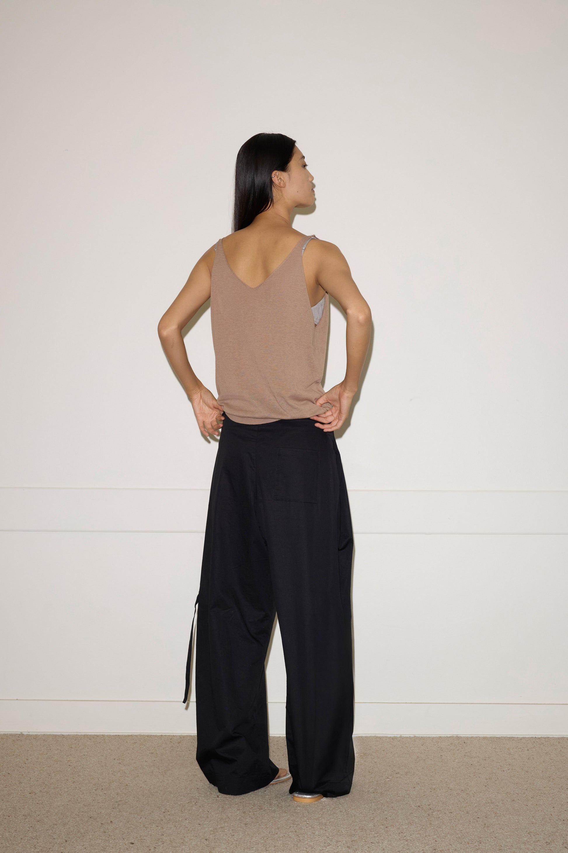 A person with long dark hair stands barefoot, facing away from the camera, wearing a brown sleeveless top and the Tab Pant in Black—loose organic cotton wide leg trousers—against a plain light-colored wall.