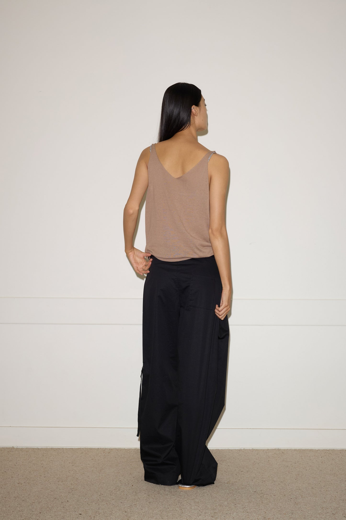 A person with long dark hair stands facing away from the camera, wearing a 0 Rolled Knit Top - Light Brown and loose black pants, against a plain white wall and carpeted floor.