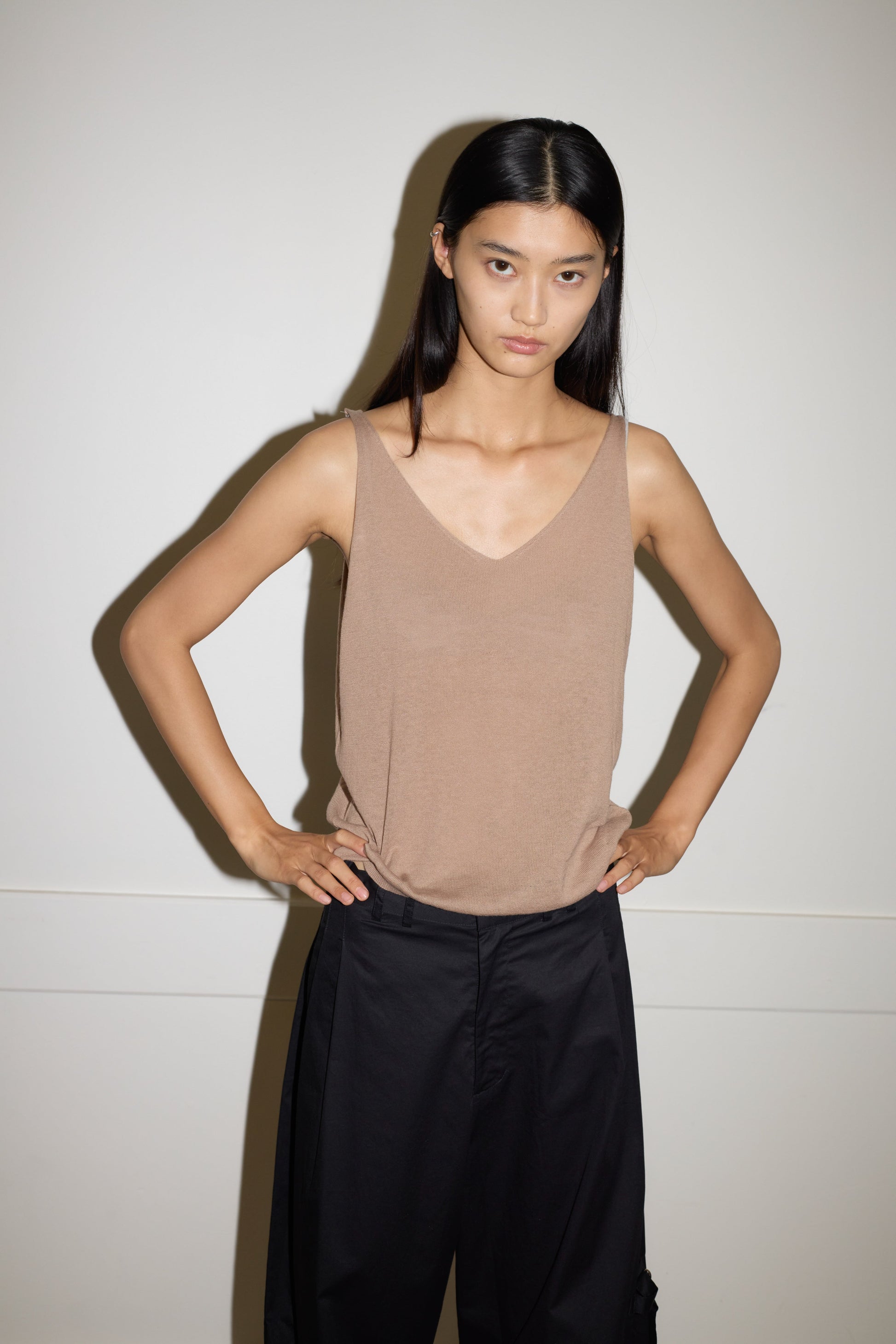 A person with long dark hair stands against a plain wall, wearing the Rolled Knit Top - Light Brown from 0 and black pants, hands on hips and a neutral expression.