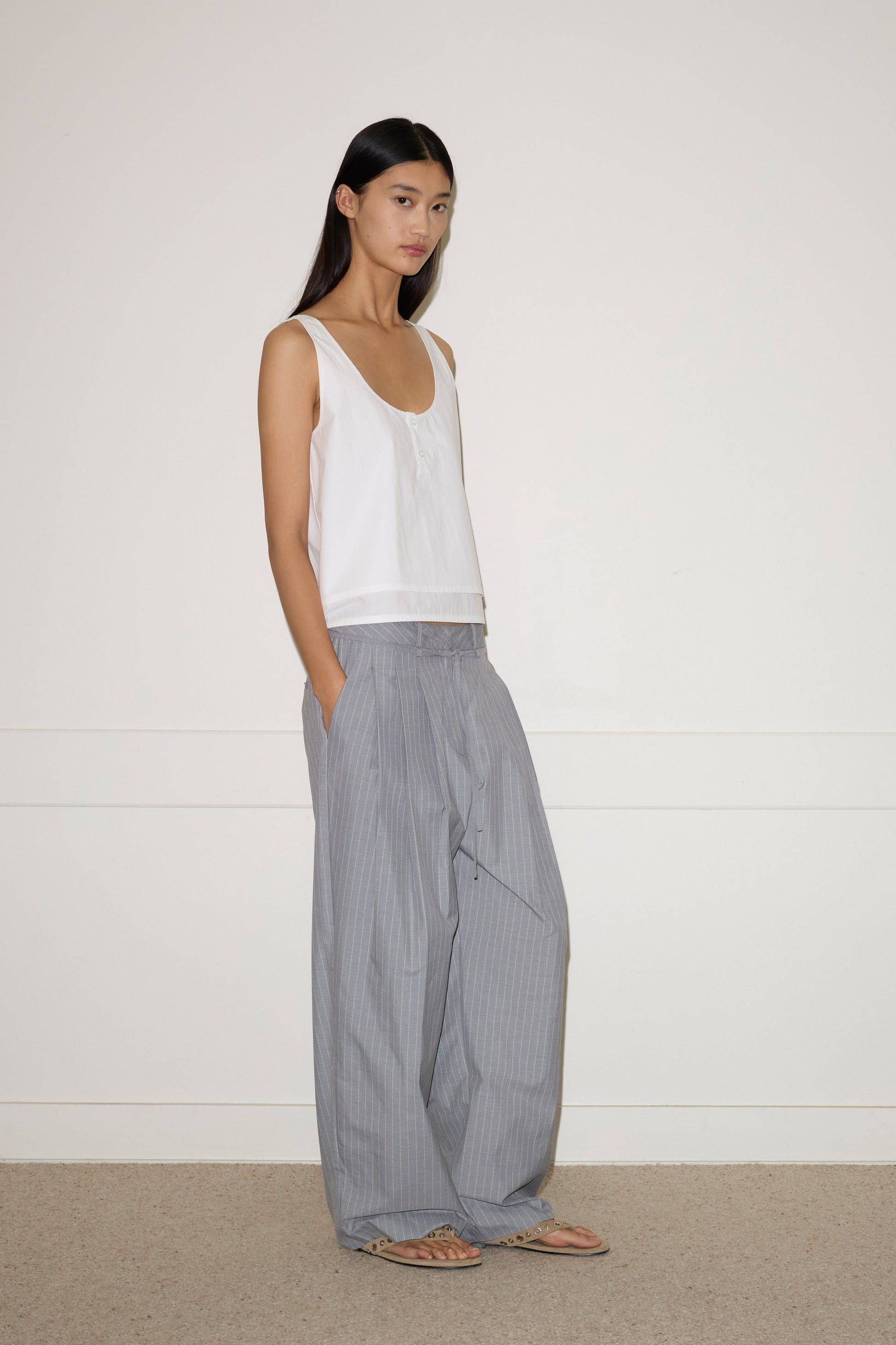 A woman with long dark hair stands against a plain wall, wearing the Layered Top - White by 0, paired with loose gray pinstriped pants and sandals. Her hands are in her pockets as she gazes slightly to the side.