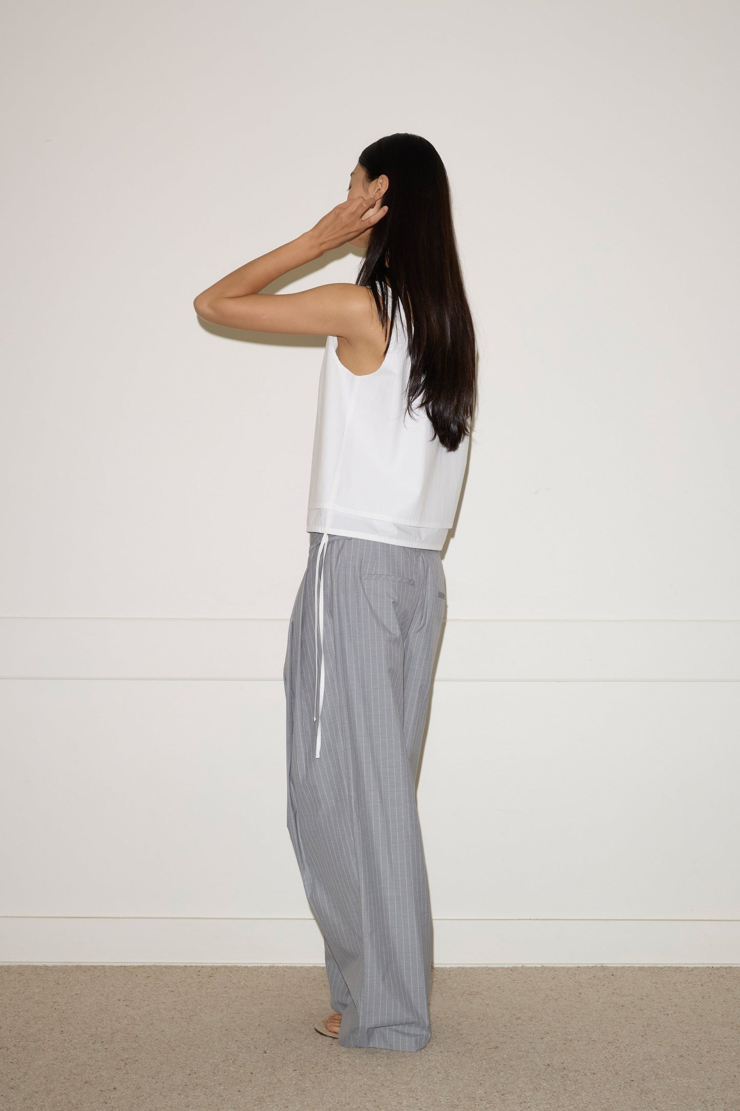 A woman with long dark hair stands sideways by a white wall, wearing a sleeveless white top and the Tri Pleat Pure Wool Pant - Steele Stripe from Deiji Studios. She raises one hand to the side of her head.