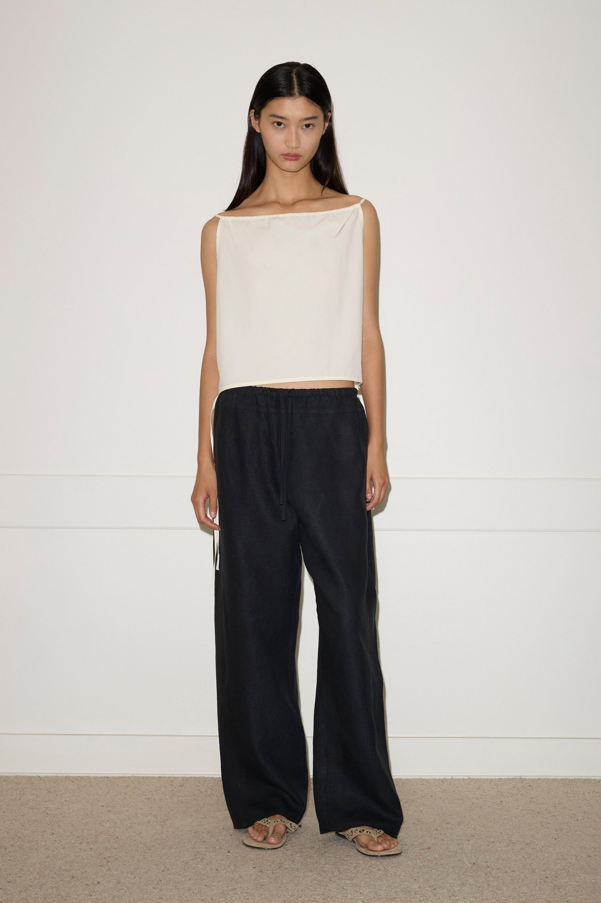 A woman with long, straight dark hair and a neutral expression stands against a white wall wearing the Double Waist Linen Pant in black, paired with a sleeveless off-white top and sandals.