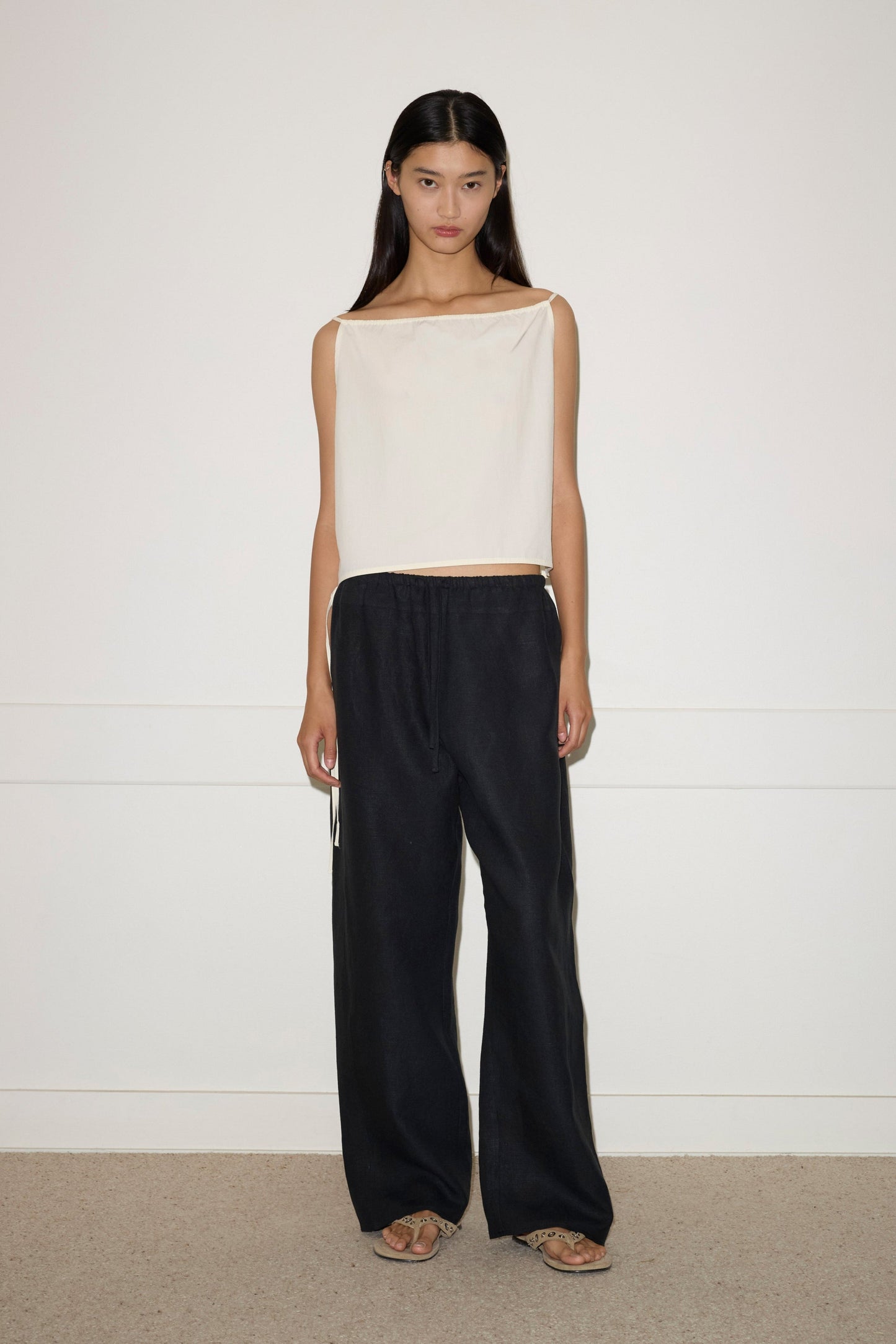 A woman with long dark hair stands indoors against a plain white wall, wearing the Simple Top, paired with loose black pants and sandals. She faces the camera with a neutral expression.