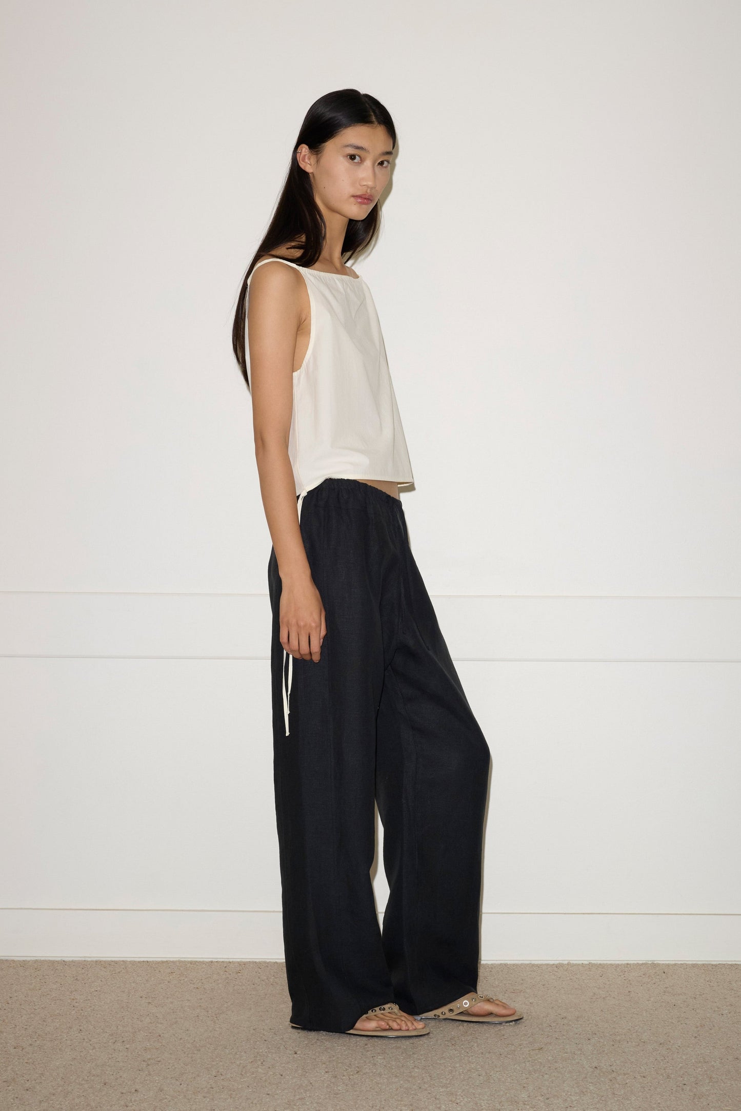 A woman with long dark hair stands sideways against a white wall, wearing a sleeveless white top, the Double Waist Linen Pant in black, and sandals. She gazes at the camera with a neutral expression.