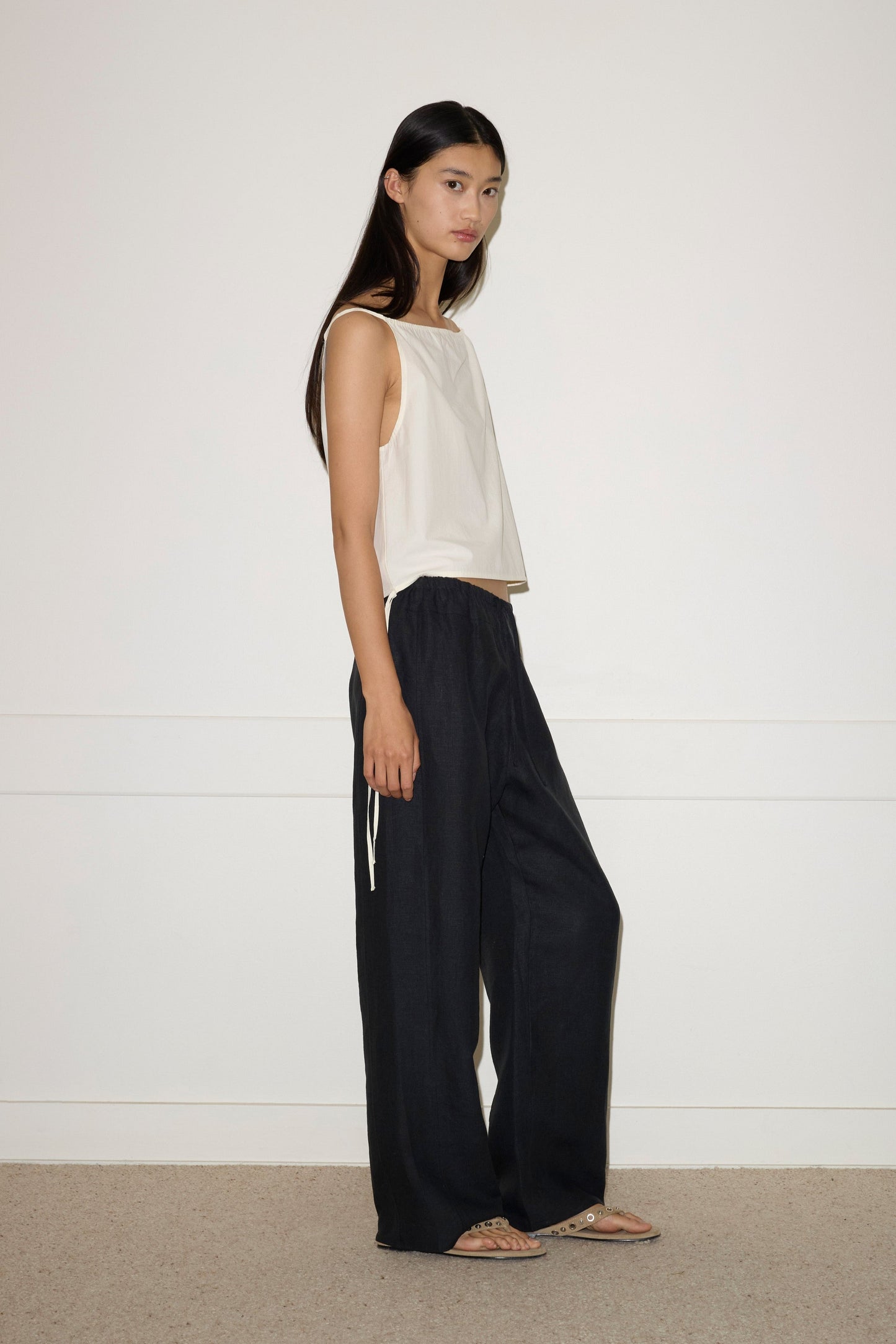 A woman with long dark hair stands sideways against a plain white wall, wearing the Simple Top - Butter White by Deiji Studios, loose black pants, and flat sandals.