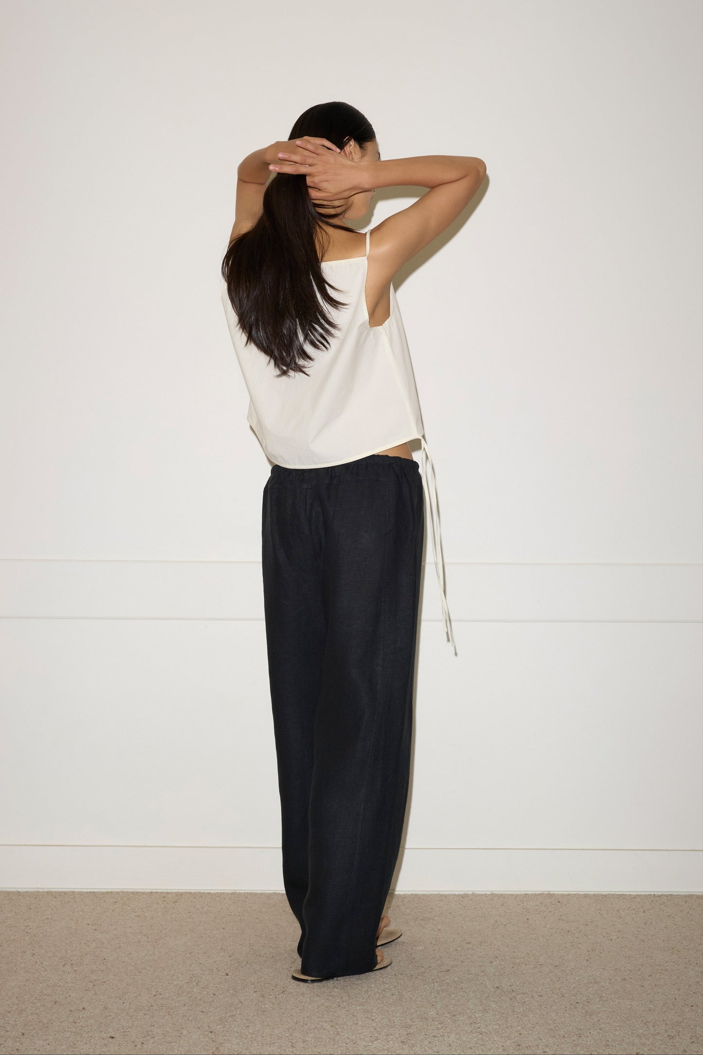 A woman with long dark hair stands facing a white wall, seen from behind. She wears a sleeveless white top and the Double Waist Linen Pant in Black, lifting her arms to adjust her hair.