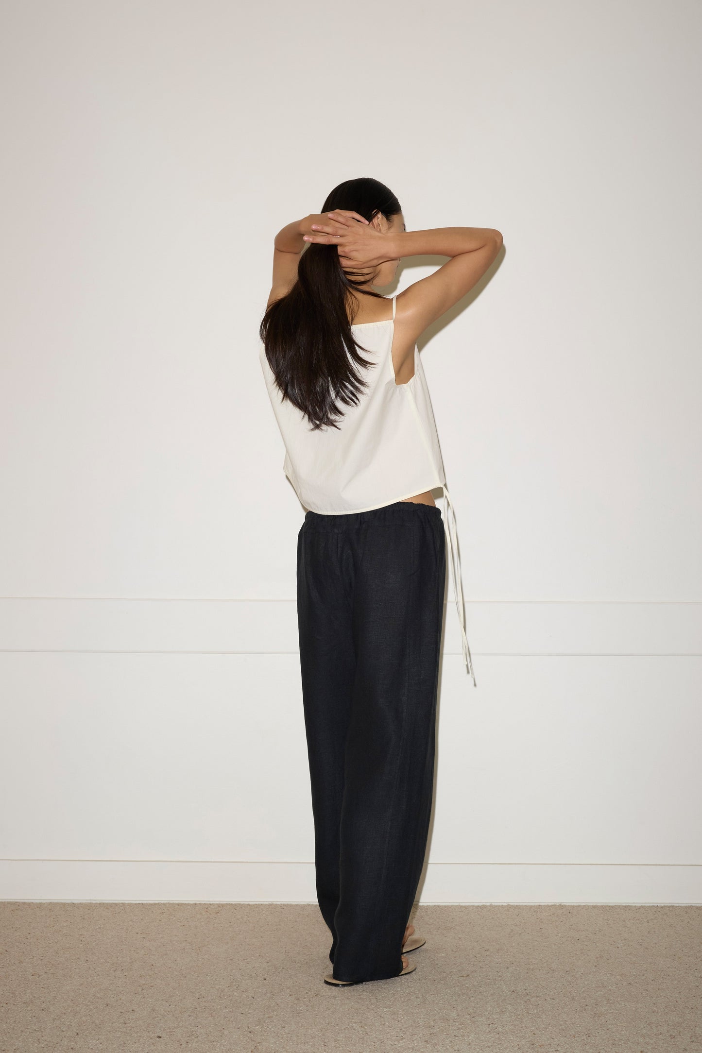 A woman stands facing a white wall with raised arms, hands resting on her head. She wears the Simple Top - Butter White from Deiji Studios and wide-leg dark pants, her long dark hair cascading down her back.