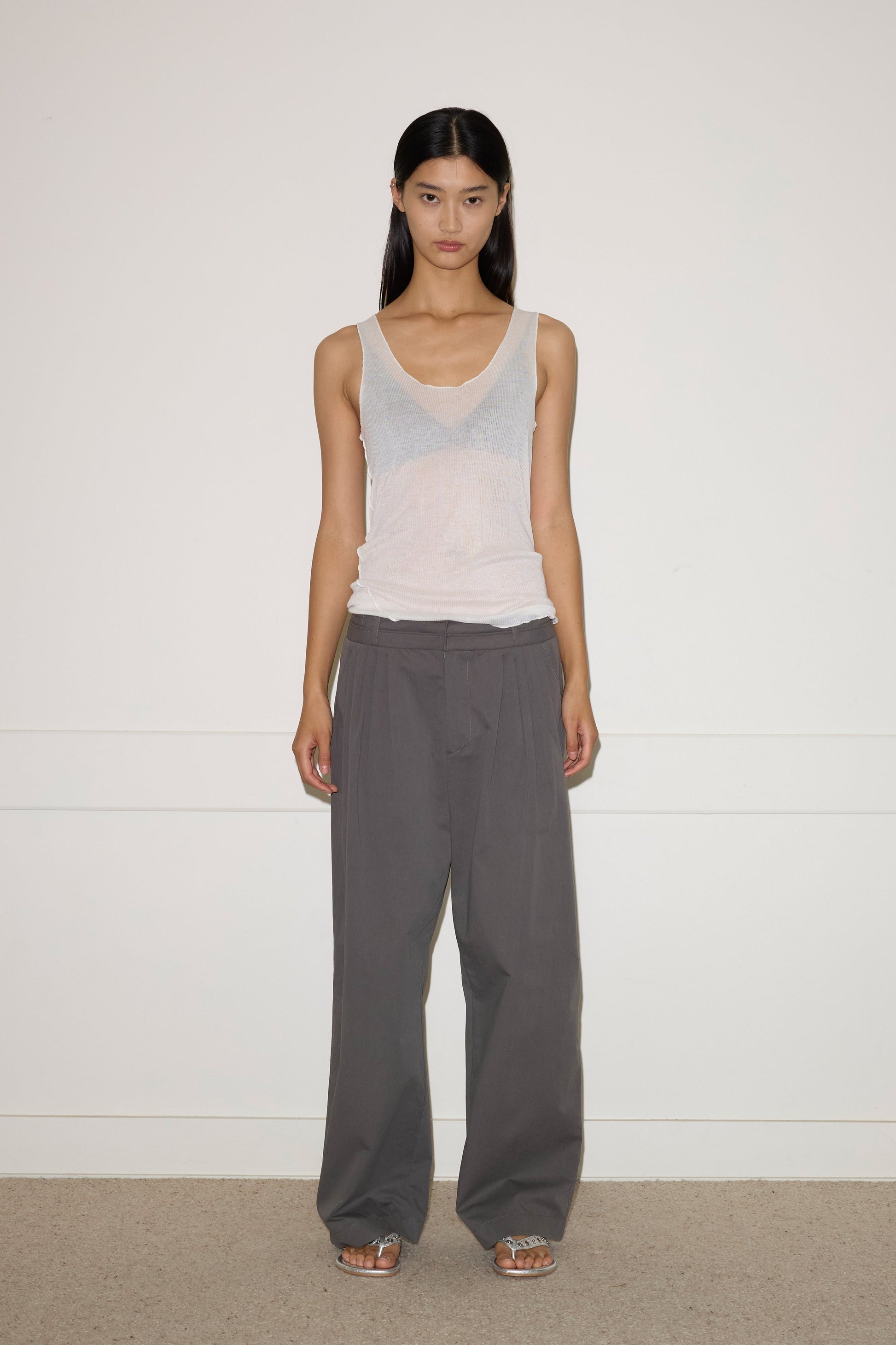 A woman with straight dark hair and a neutral expression stands against a white wall, wearing the 0 Two Layer Pant in Charcoal, a sheer white tank over a light bra, and silver open-toe sandals.