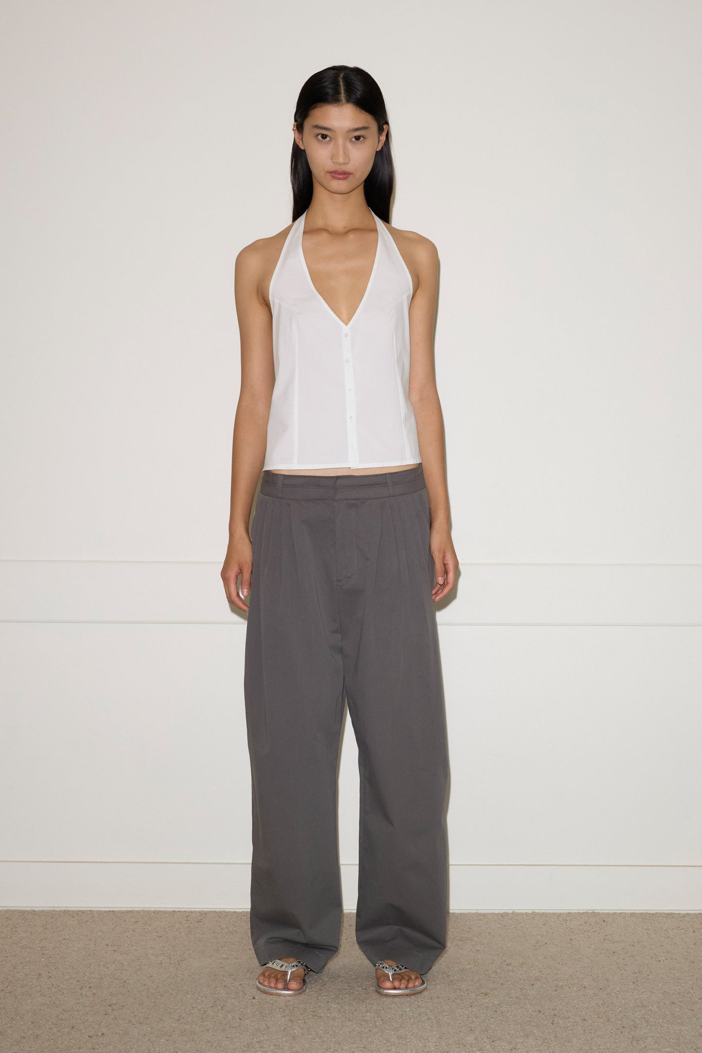 A woman with straight dark hair stands against a plain white wall, wearing Deiji Studios' Waist Halter - White with a deep V-neck, loose gray trousers, and flat sandals.