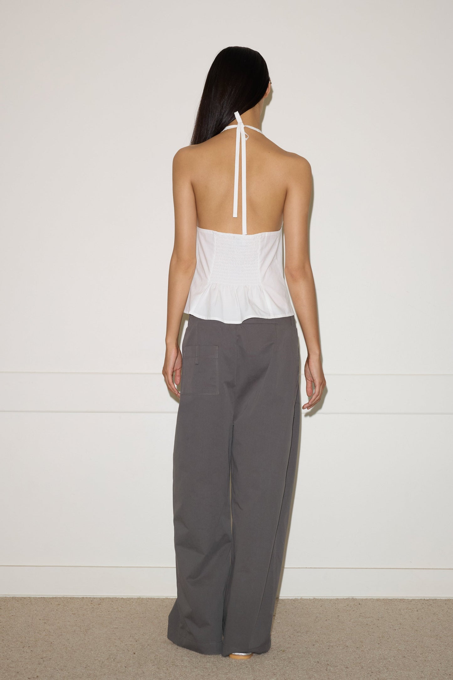 A person with long dark hair faces a white wall, wearing a white halter top and the 0 Two Layer Pant - Charcoal, made from gray organic cotton. The minimalist indoor setting emphasizes the relaxed, effortless look.