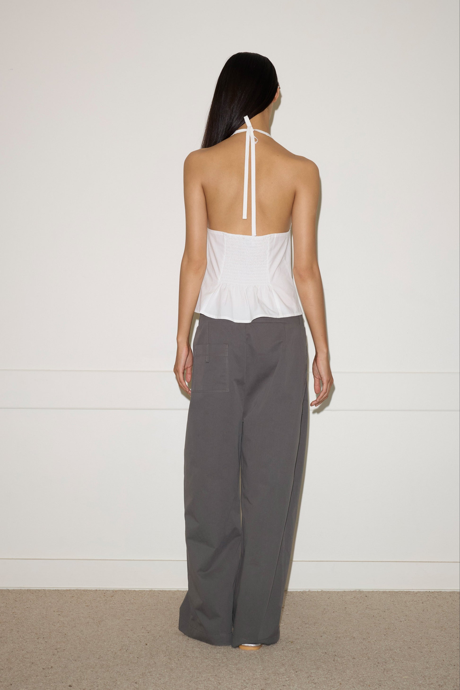 A person with long dark hair faces a white wall, wearing the Waist Halter - White by Deiji Studios and loose gray pants. The simple indoor setting features carpeted flooring.