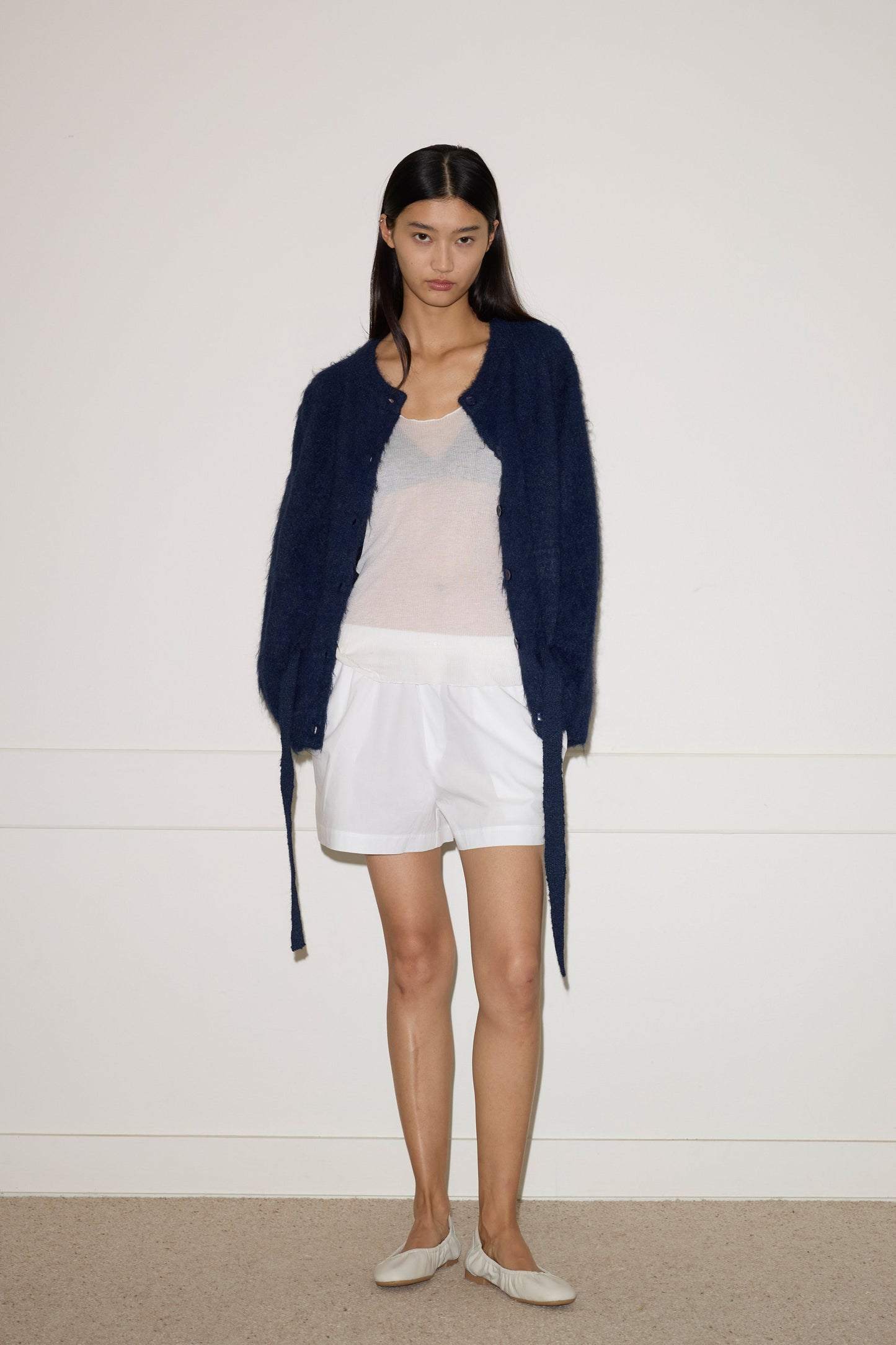 A woman with long, straight dark hair and a neutral expression models the 0 Belted Cardi - Navy over a sheer white top, white shorts, and beige flats against a plain white background.