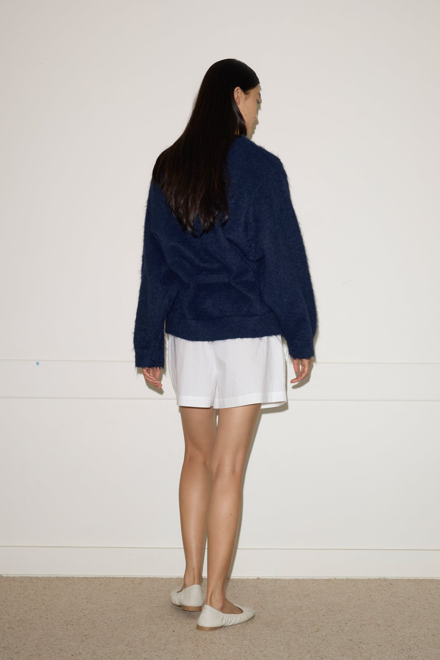 A woman with long dark hair stands facing a plain white wall, wearing the Belted Cardi - Navy by 0, paired with white shorts and slip-on shoes on a light carpet.