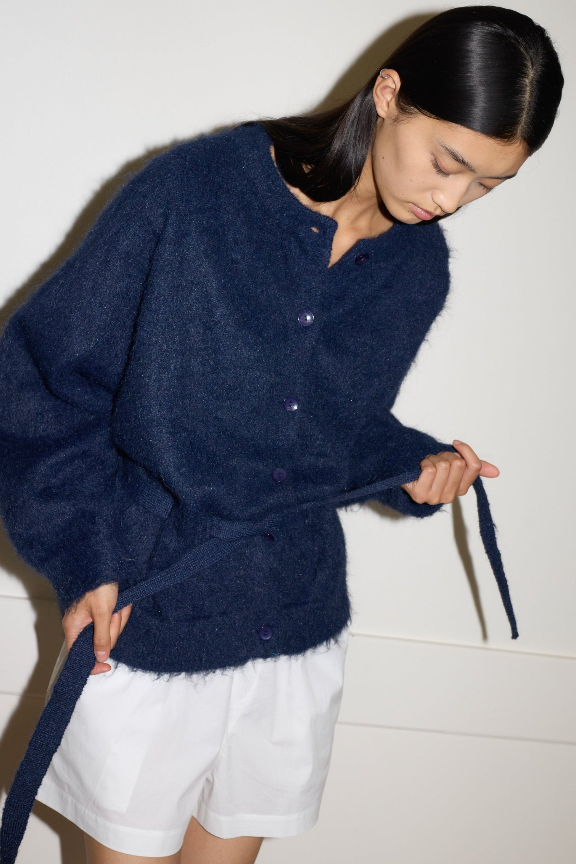 Wearing the Belted Cardi - Navy, a person with long black hair stands against a plain light wall, looking down and holding the matching belt, layering the oversized cardigan over a white skirt or shorts.