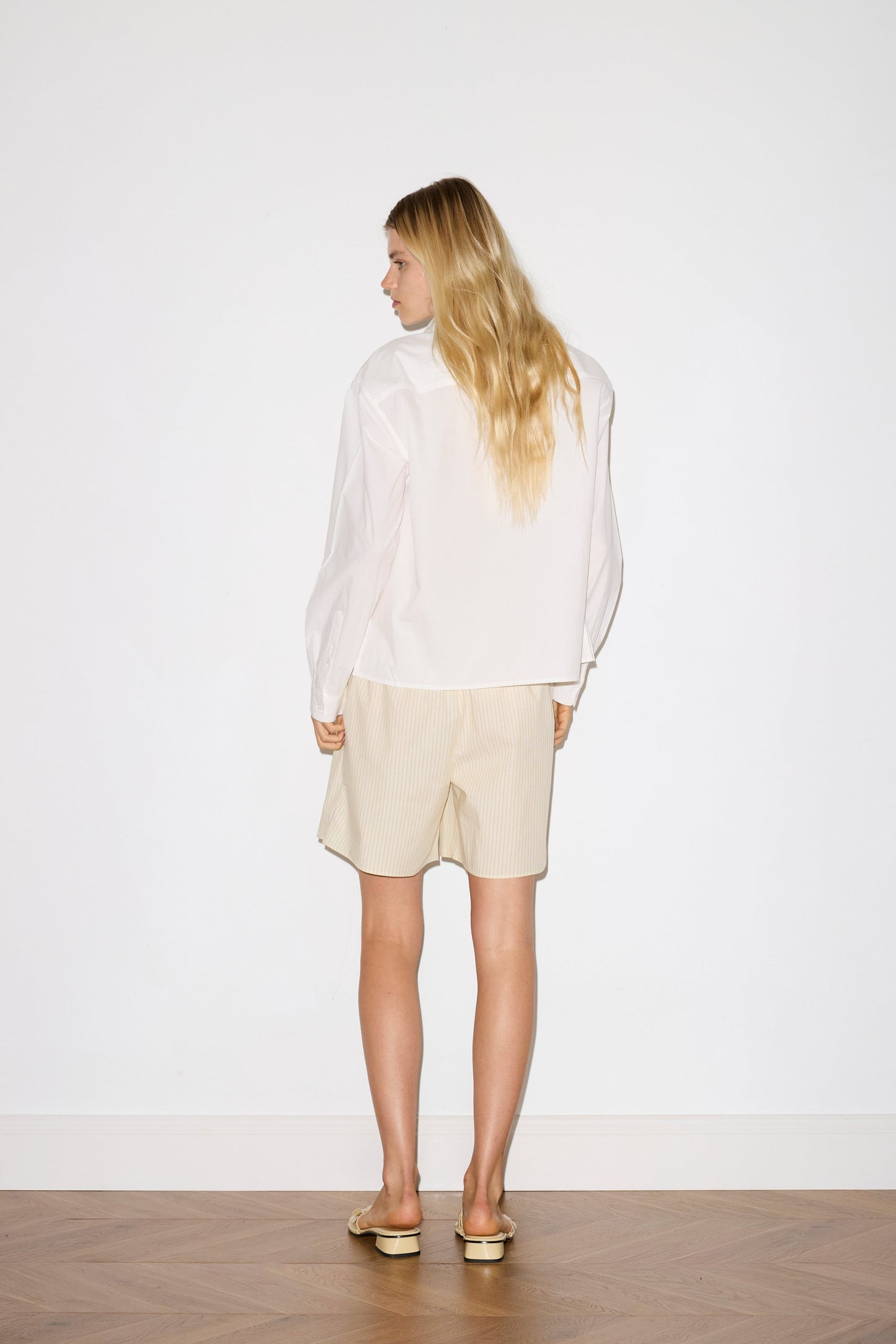blonde female model wears a white classic crisp button up shirt in a boxy fit, featuring a dropped back yoke, dropped shoulders, a classic shirting cuff with a layered arch fabric detail on front with tie, paired with a butter yellow striped easy pull-on boxer featuring button detail at front placket, gathered elastic waistline and deep side pockets in mid length for a relaxed fit
