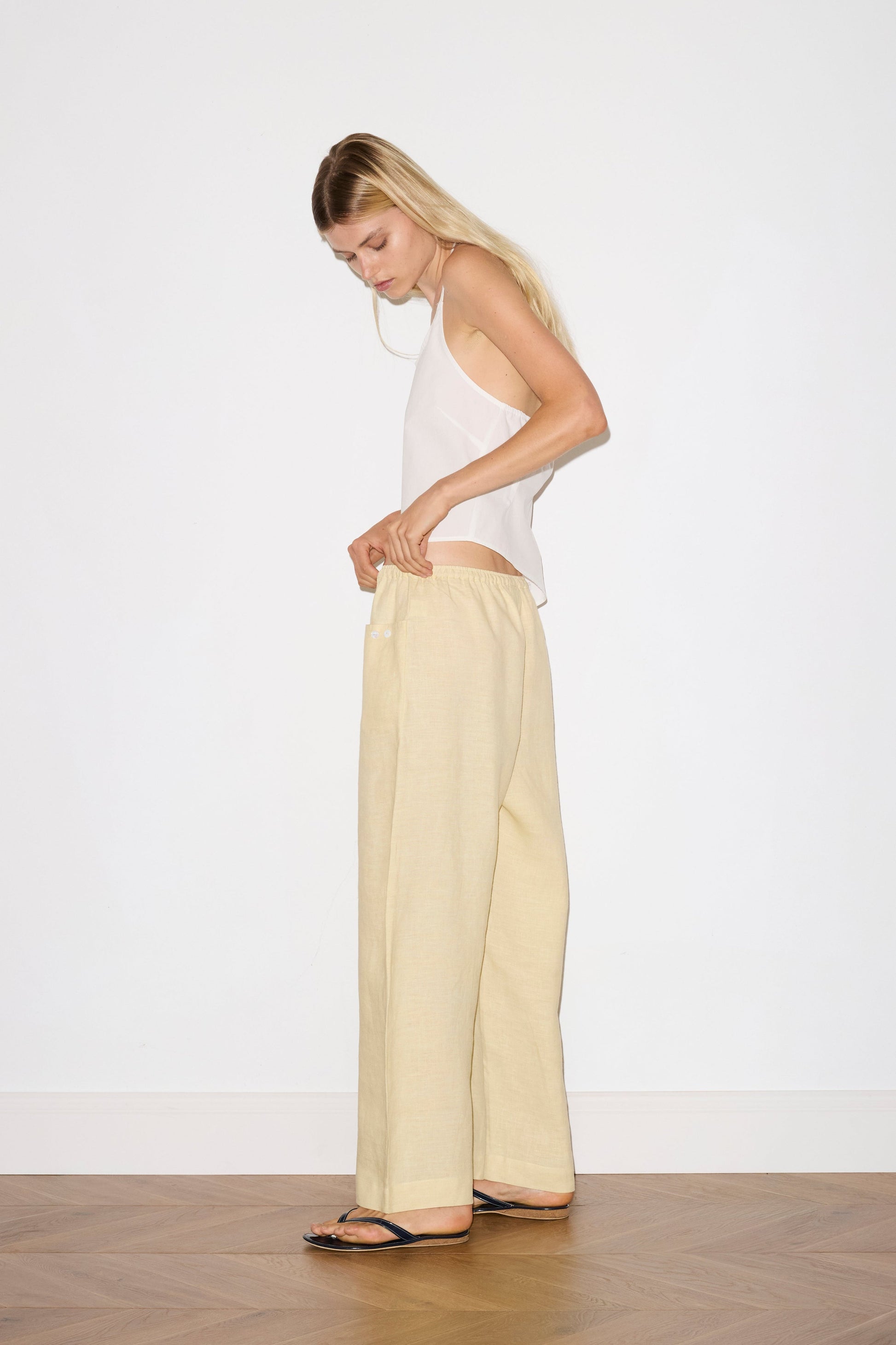 blonde haired female model wears a butter yellow ankle length, straight leg pant features a soft minimal elastic waist with front seam pockets and button detail, paired with a white relaxed halter top features flattering curved neckline, asymmetrical bottom hem feature with side tie opening, keyhole with button closure at side and fine tie halter straps.