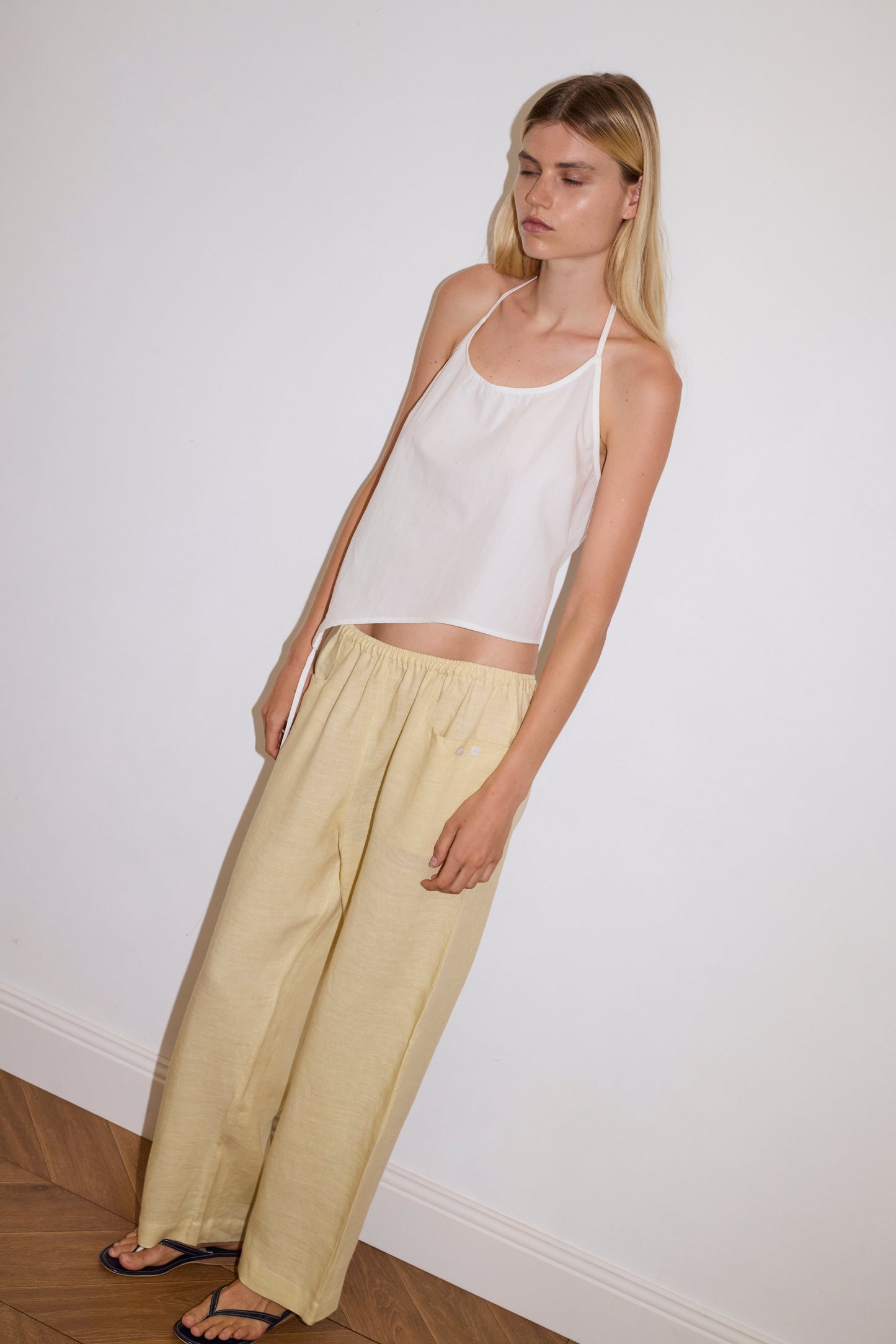blonde haired female model wears a butter yellow ankle length, straight leg pant features a soft minimal elastic waist with front seam pockets and button detail, paired with a white relaxed halter top features flattering curved neckline, asymmetrical bottom hem feature with side tie opening, keyhole with button closure at side and fine tie halter straps.